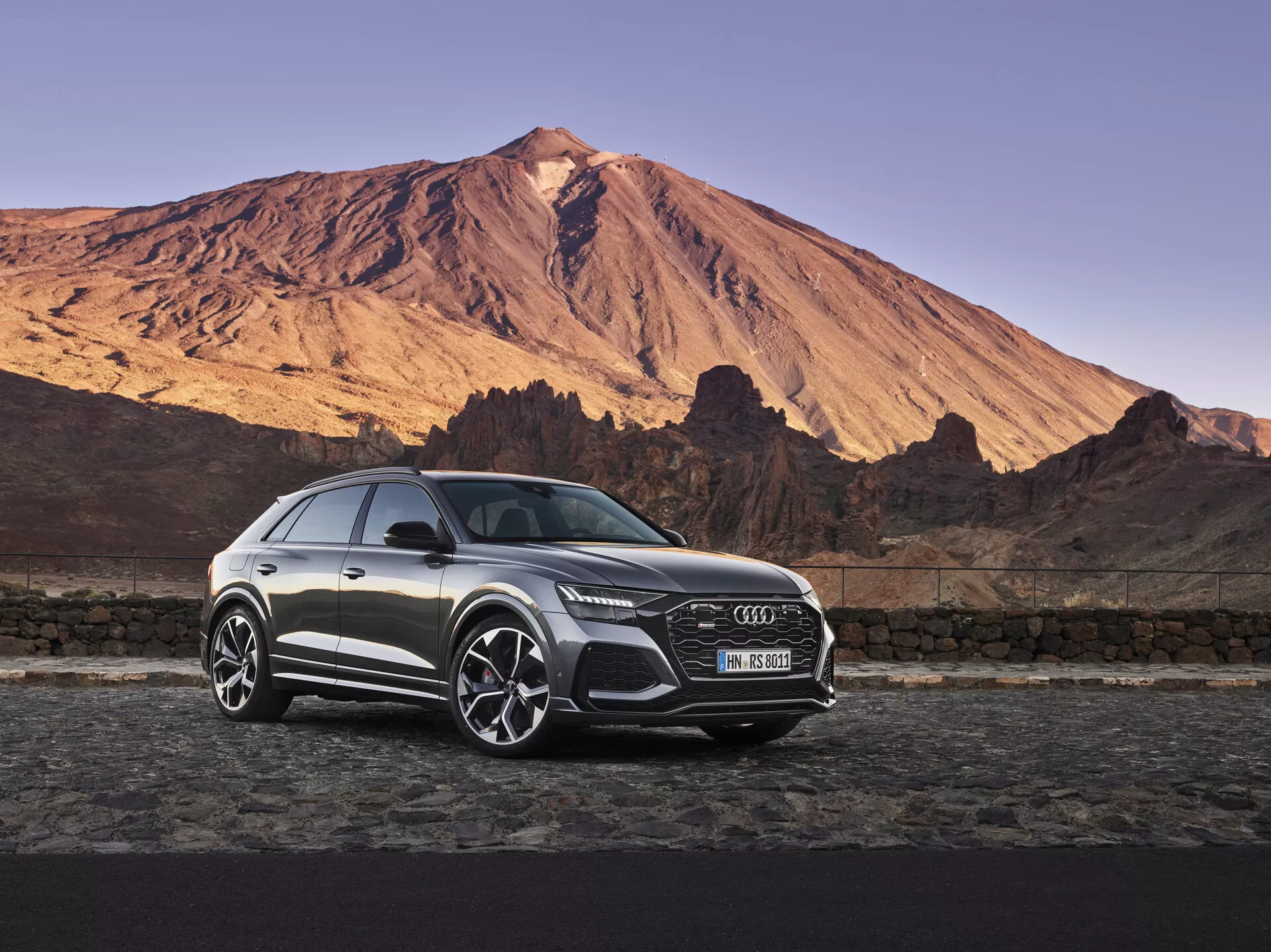 Lussuoso SUV Audi Q8 2018 parcheggiato contro un drammatico sfondo montuoso all'alba o al tramonto sotto un cielo limpido, mostrando la prominente griglia anteriore, fari a LED, cerchi in lega e caratteristiche di design elegante in una morbida illuminazione naturale.