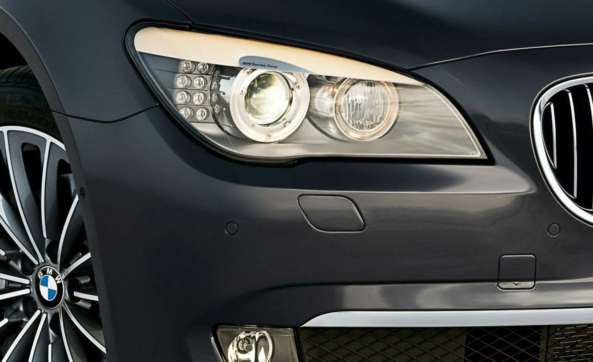 Vista frontale laterale di una BMW Serie 7 del 2005 con emblema della marca, gruppo ottico moderno con lente a proiettore e luci diurne LED, vista parziale della griglia a rene e ruota in lega a più razze.