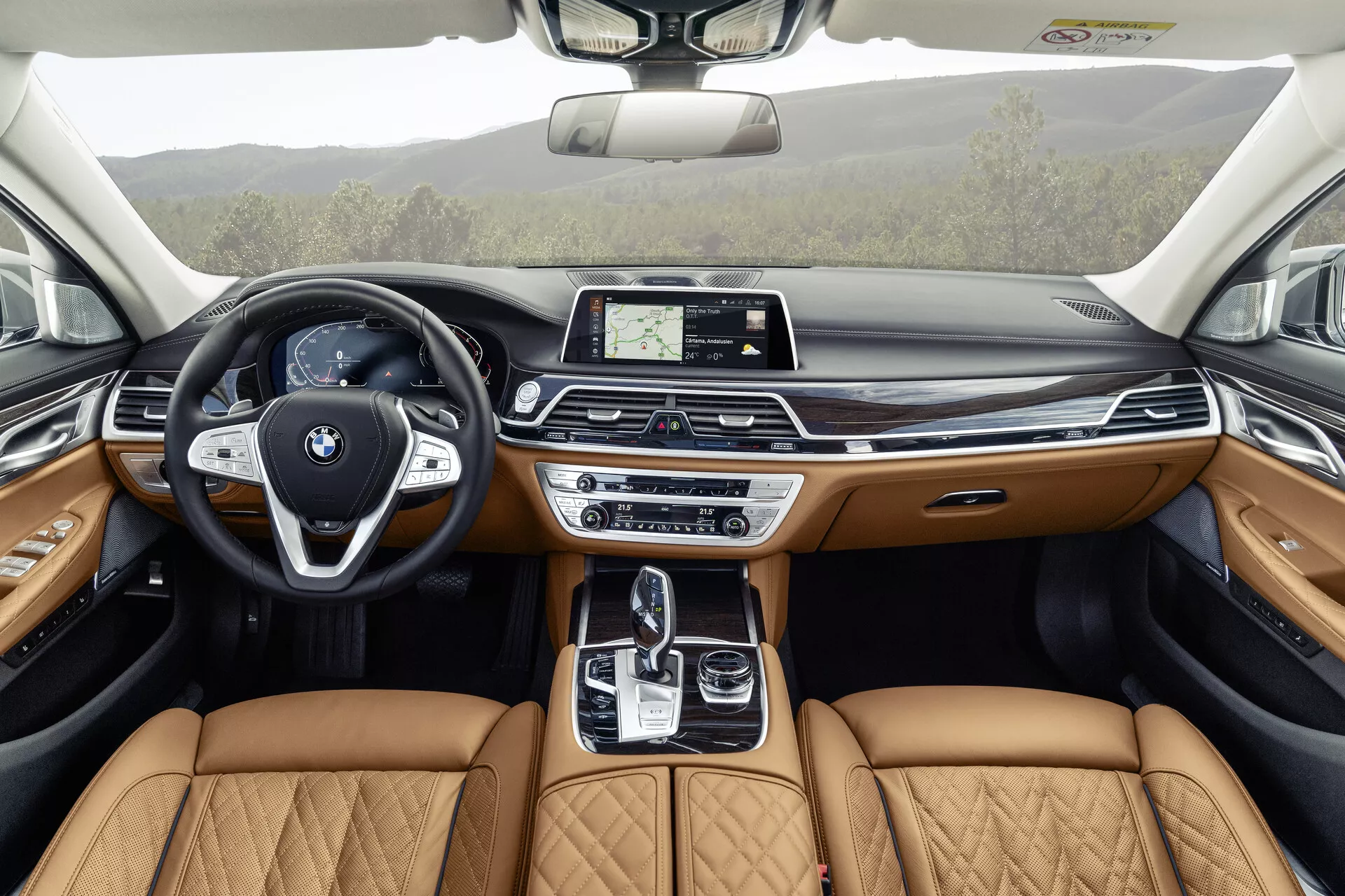 L'immagine mostra l'interno di una BMW Serie 7 del 2019 (restyling G11/G12) vista dalla prospettiva del sedile posteriore, evidenziando il design dettagliato e lussuoso dell'abitacolo anteriore. Dotato di un volante moderno con tasti multifunzione e un distintivo emblema BMW, il cruscotto elegante ospita un display centrale touchscreen per l'infotainment. La consolle centrale include una leva del cambio automatico, un touchpad e vari sistemi di controllo dinamico, completati da sedili in pelle trapuntata beige che indicano un allestimento di livello premium. I pannelli delle porte riflettono questo lusso con pelle beige, finiture in legno e illuminazione ambientale, suggerendo che caratteristiche avanzate di tecnologia e comfort sono integrate in tutto il veicolo, incluso un tetto apribile per una maggiore luce naturale.