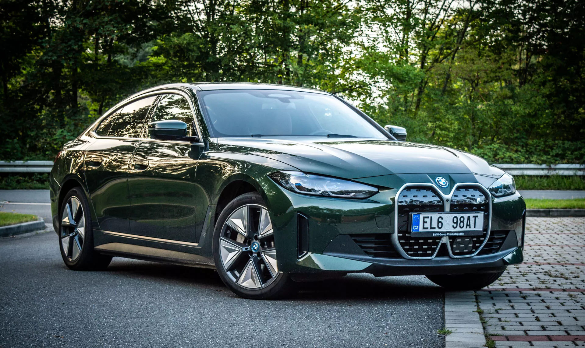 Berlina BMW i4 verde a quattro porte del 2021 parcheggiata su una strada in mattoni accanto a un'area erbosa con fogliame sullo sfondo, vista frontale che mette in evidenza la grande griglia, i fari LED, le prese d'aria e i cerchi in lega