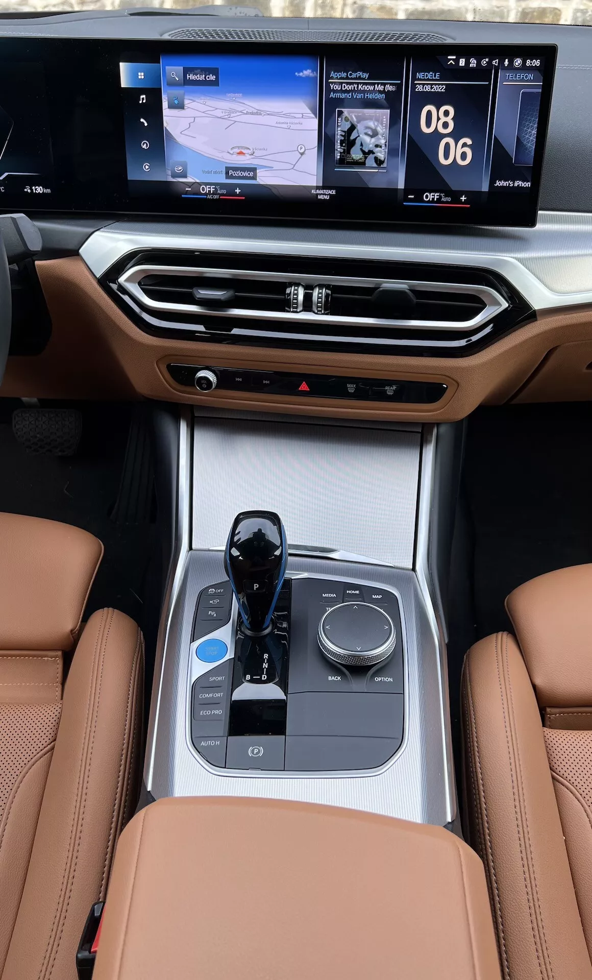 Vista della consolle centrale interna di una BMW i4 2021 dai sedili posteriori, mettendo in evidenza i sedili in pelle, il selettore di marcia elettronico, la manopola di controllo multimediale, i portabicchieri, i controlli del clima e lo schermo dell'infotainment a doppia interfaccia con navigazione e display dei media