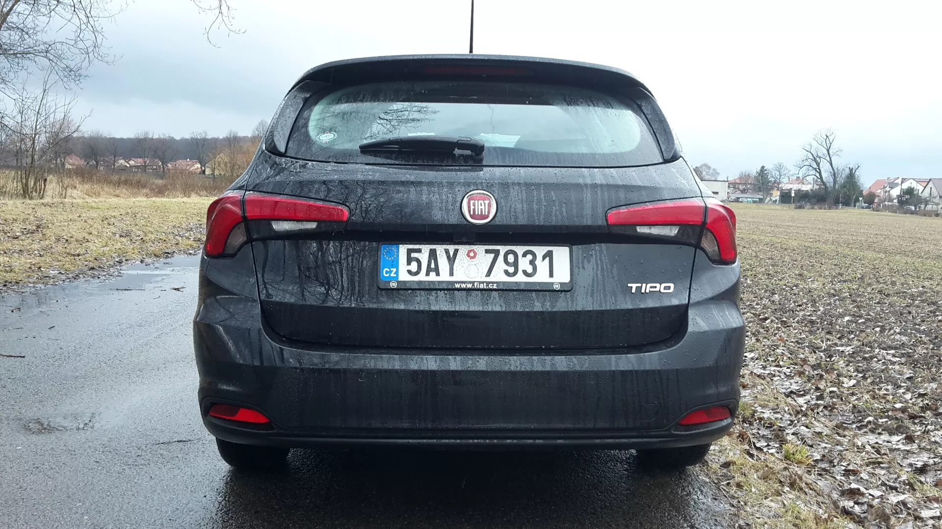 Vista posteriore di una berlina Fiat Tipo 2016 sotto cieli nuvolosi, con identificazione di marca e modello sul bagagliaio, luci posteriori e terza luce di stop dettagliate, targa e un accenno di ambiente rurale sullo sfondo