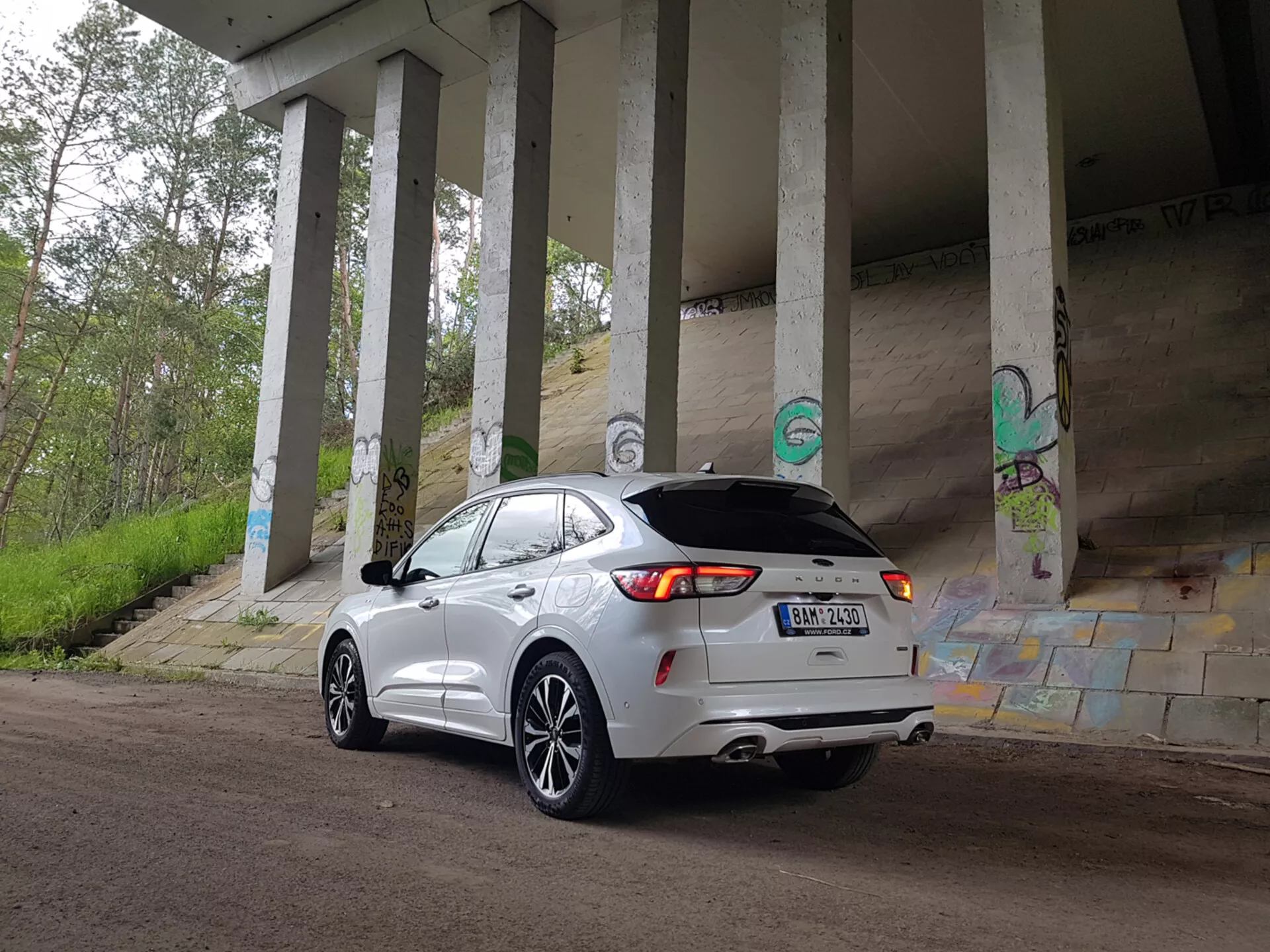 SUV crossover Ford Kuga 2019 parcheggiato sotto un cavalcavia, che mostra il suo design moderno e le caratteristiche tecnologiche come le luci posteriori a LED, il doppio scarico e la telecamera posteriore, tra graffiti urbani e pilastri di cemento