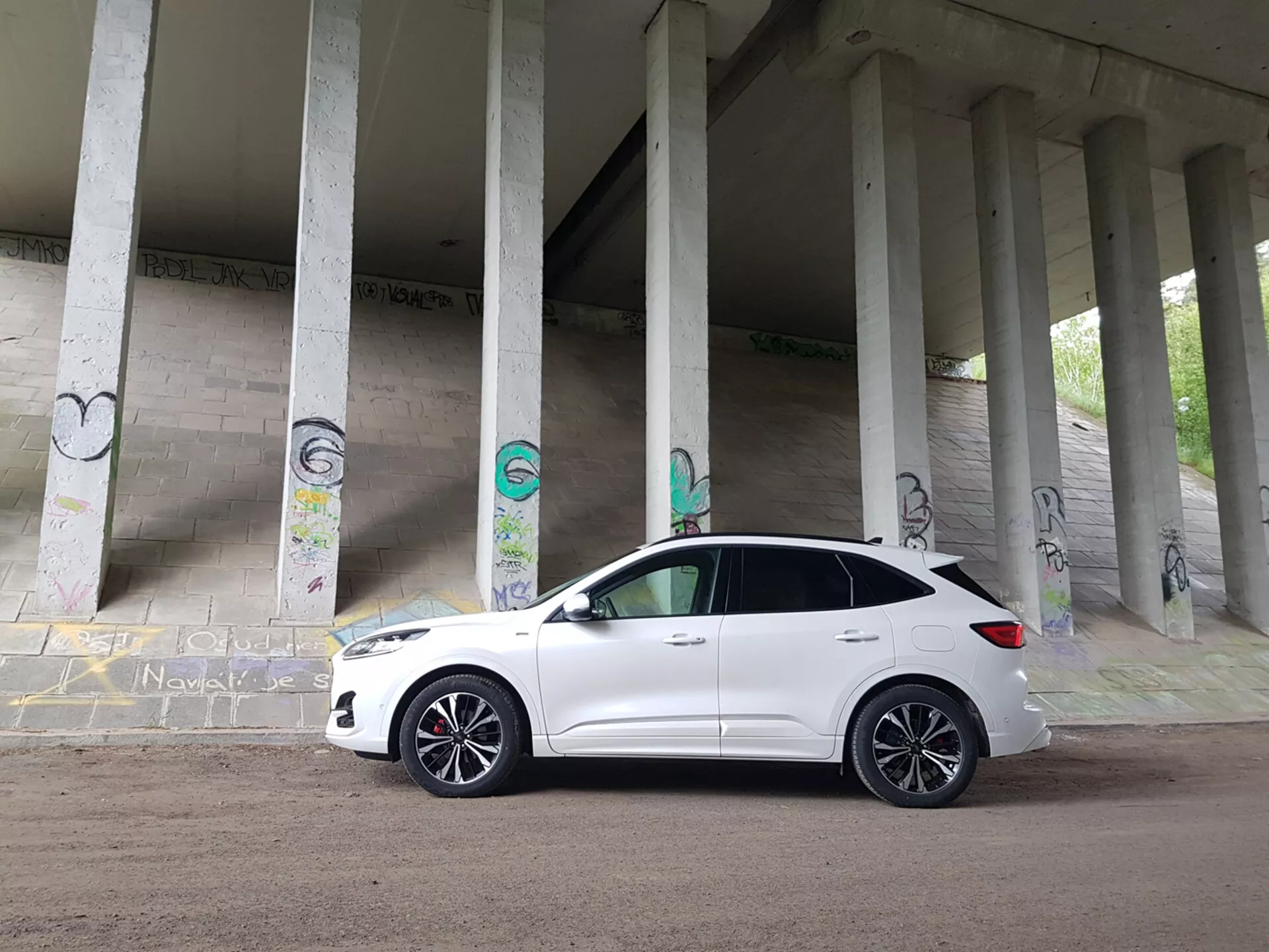 SUV compatto Ford Kuga 2019 bianco parcheggiato sotto una struttura con pilastri di cemento coperti di graffiti, mostrando il suo profilo dal quarto anteriore includendo la griglia, i fari e le ruote in lega