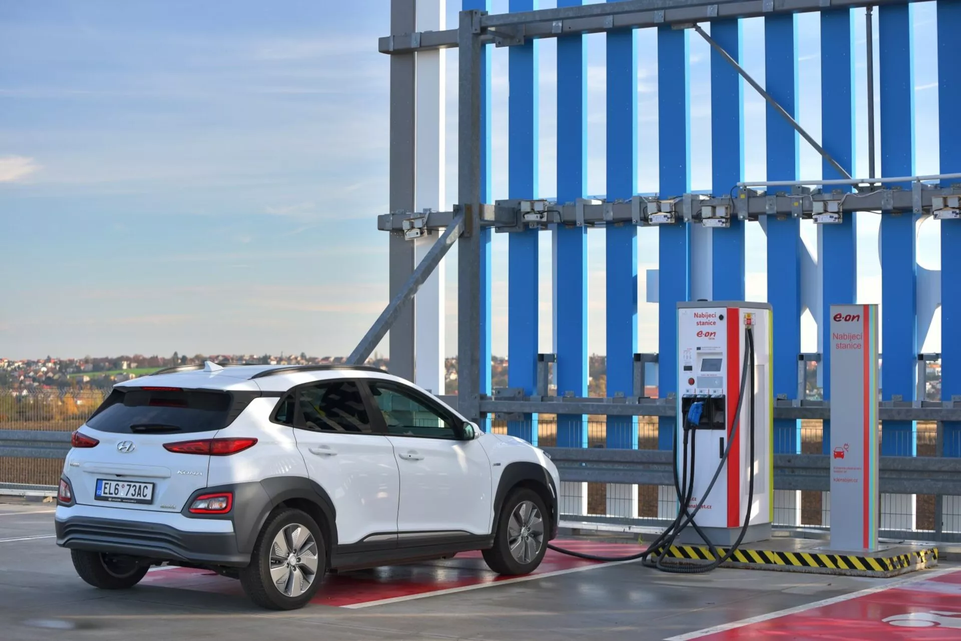 SUV elettrico Hyundai Kona 2021 in bianco, mostrando la sua visuale posteriore da tre quarti presso una stazione di ricarica all'aperto con cielo sereno sullo sfondo