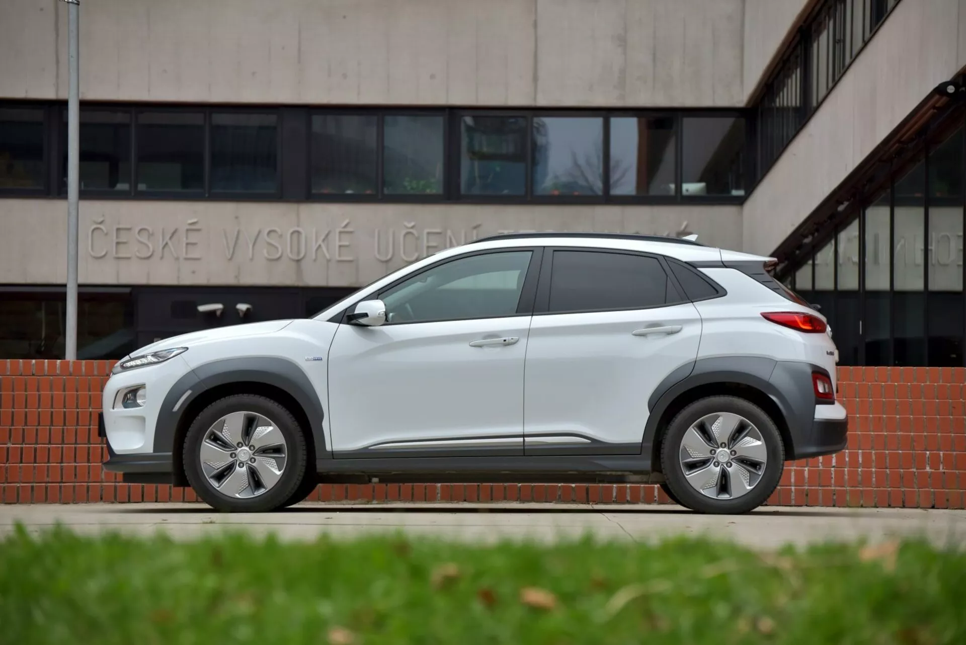 Vista laterale del Hyundai Kona Elektro 2021 in grigio chiaro, evidenziando il suo design elegante con passaruota e minigonne dello stesso colore della carrozzeria e funzionalità moderne come cerchi in lega e antenna a pinna di squalo, davanti a un edificio con il testo 'ČESKÉ VYSOKÉ UČENÍ', enfatizzando la sua idoneità per la guida urbana e fuoristrada
