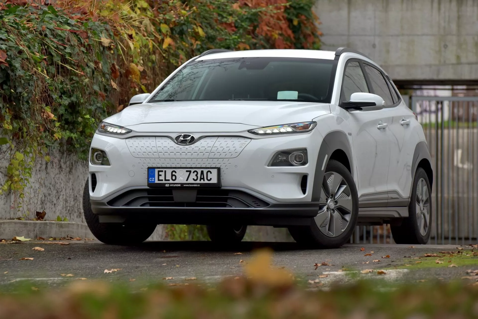 SUV compatto elettrico Hyundai Kona 2021 parcheggiato sul bordo della strada urbana, mostrando griglia anteriore, fari LED eleganti, ruote in lega e design del corpo scolpito con enfasi sul lato anteriore sinistro, in mezzo a un paesaggio autunnale