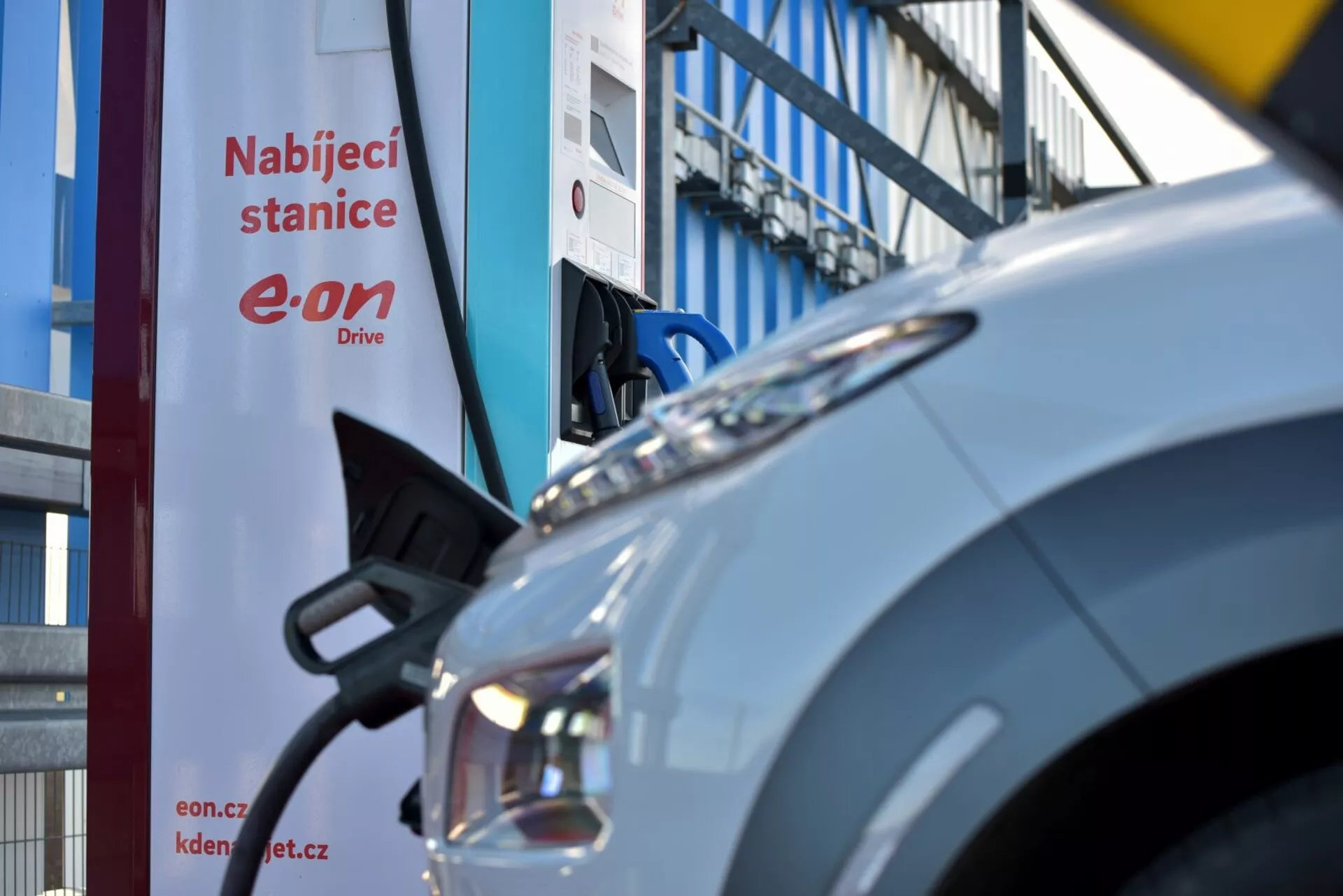 Hyundai Kona Elettrica 2021 parcheggiata presso una stazione di ricarica, che mostra il suo pannello anteriore, gruppo faro e porta di ricarica, con un cartello 'Nabíjecí stanice' parzialmente visibile