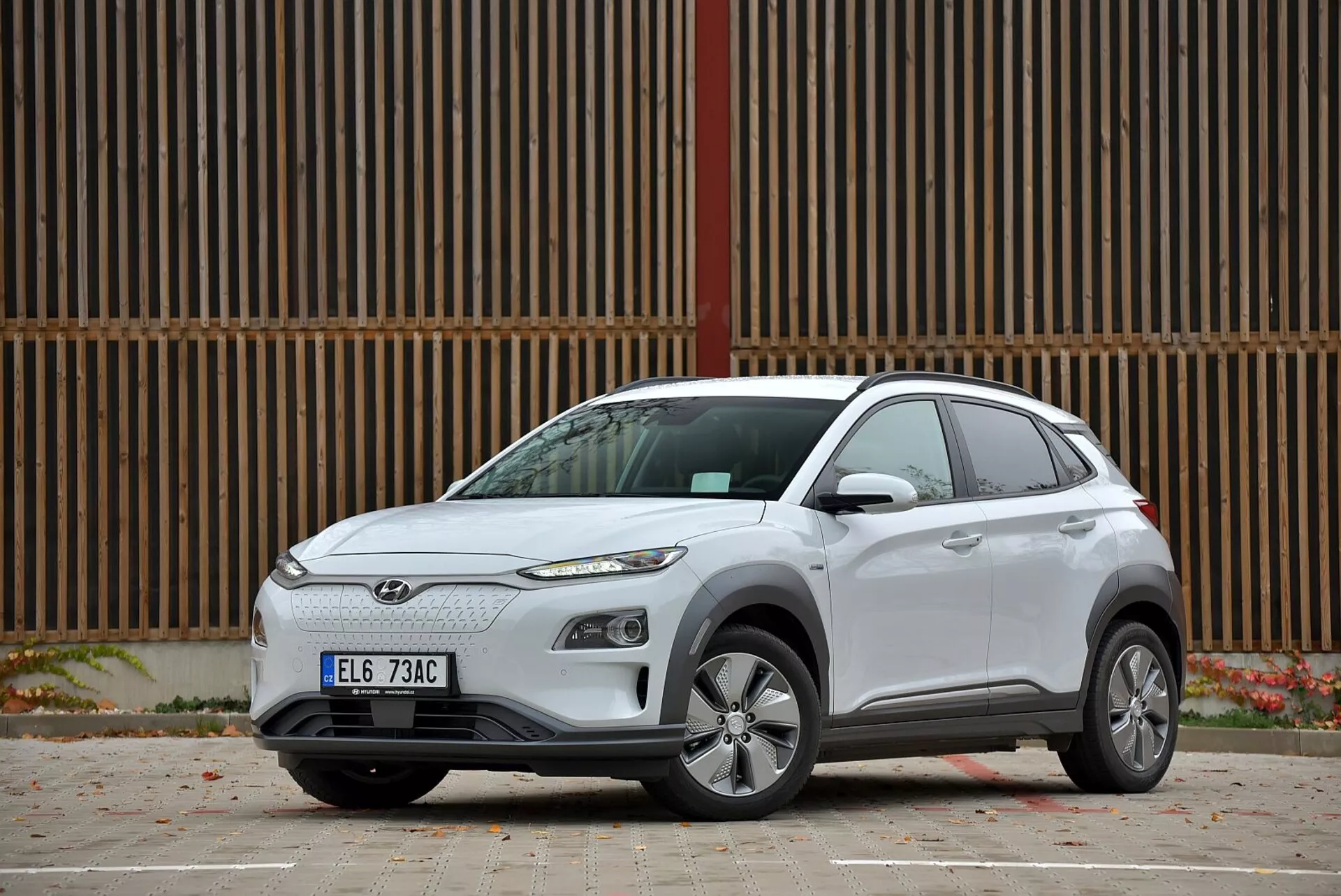 SUV compatto elettrico Hyundai Kona del 2021 in bianco, mostra il suo design moderno da un profilo laterale a tre quarti, con griglia per lo più chiusa, fari taglienti, ruote in lega, indicatori di direzione integrati su specchietti laterali del colore della carrozzeria, maniglie delle porte standard, vetri scuri per le finestre posteriori, accenno di spoiler posteriore, parcheggiata su una superficie pavimentata in un contesto urbano.