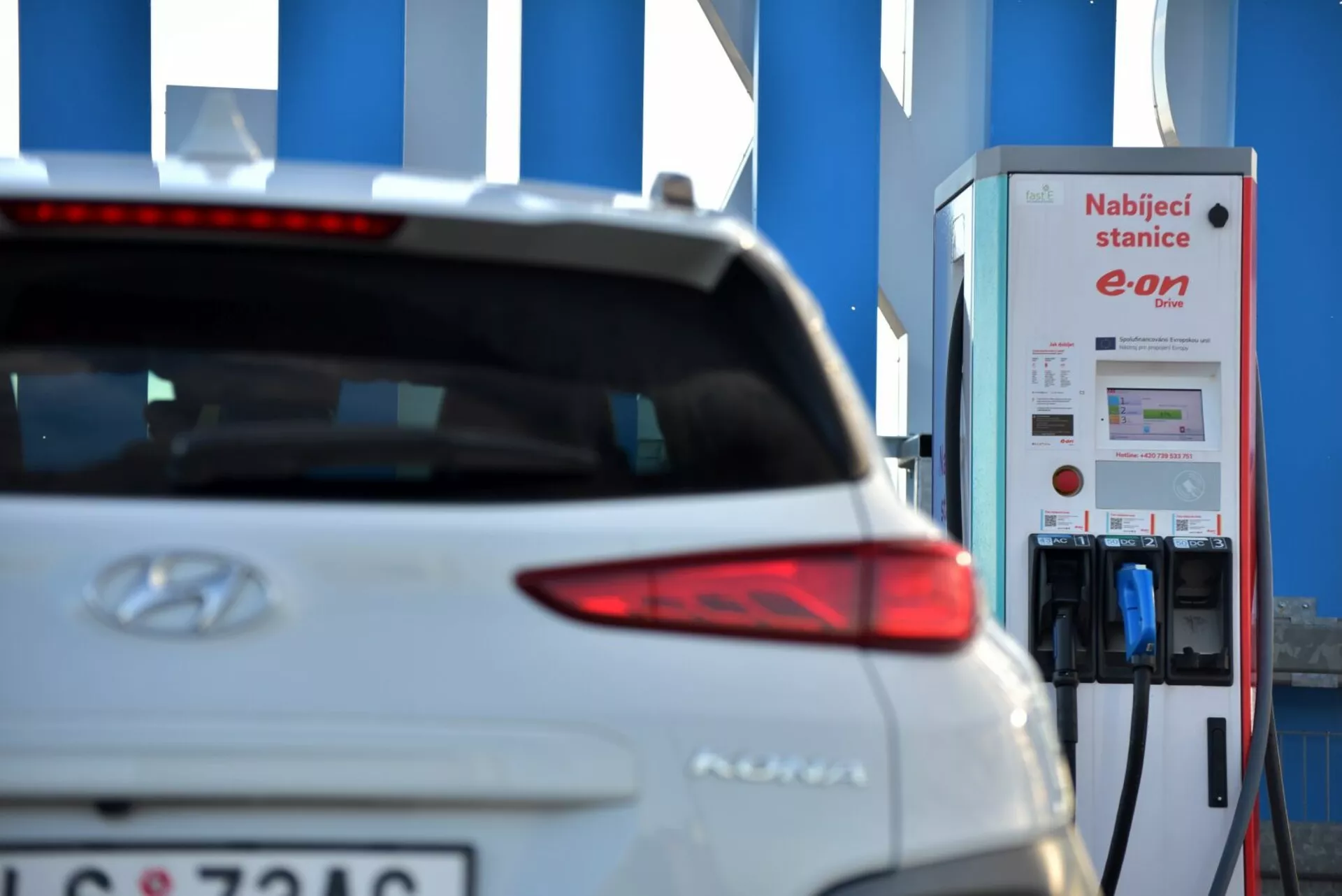 Immagine ravvicinata della sezione posteriore destra di un veicolo elettrico Hyundai Kona 2021 in bianco, concentrandosi sul design elegante della luce di coda, con il logo Hyundai leggermente visibile e il nome del modello vicino al centro della porta del bagagliaio. La targa parzialmente visibile nella parte inferiore sinistra della porta del bagagliaio suggerisce un'attenta attenzione alla privacy. Lo sfondo presenta una stazione di ricarica per veicoli elettrici E.ON, evidenziando la natura elettrica dell'auto in mezzo a un'impostazione sfocata, accennando alla mobilità elettrica e alla tecnologia del trasporto sostenibile.