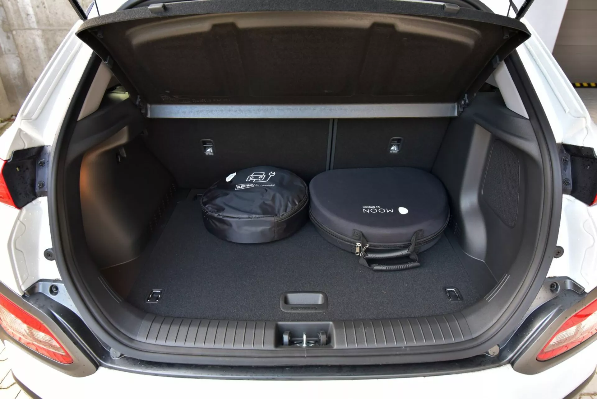 Modello elettrico Hyundai Kona 2021 che mostra lo spazio del bagagliaio da una prospettiva posteriore con il bagagliaio aperto, rivelando un'area di carico moquettata con tracce antiscivolo e oggetti minimi per una visione chiara delle dimensioni dello spazio.