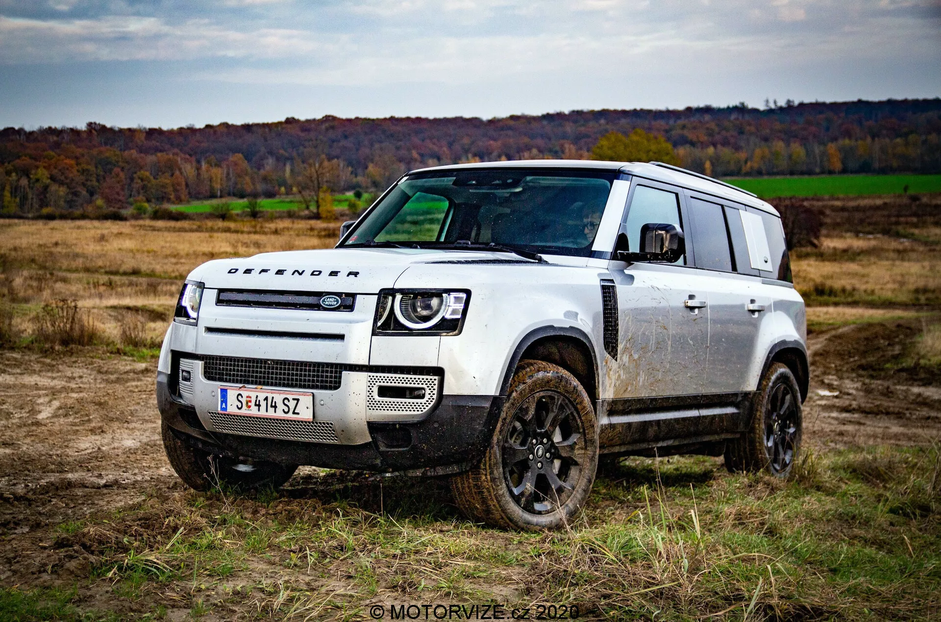 Immagine di un veicolo fuoristrada Land Rover Defender 2020 da un'angolazione di tre quarti frontale in un campo aperto con erba e alberi durante l'autunno. Il design robusto e squadrato del veicolo, ricoperto di macchie di fango, mette in evidenza le sue capacità fuoristradali. Le caratteristiche notevoli includono le ruote scure, la scritta 'DEFENDER' sul cofano, una griglia anteriore minimalista con il logo Land Rover, fari LED moderni e un robusto paraurti anteriore con piastre di protezione. Il tetto gradinato con finestre alpine e i grandi finestrini posteriori aggiungono alla funzionalità estetica dell'auto.