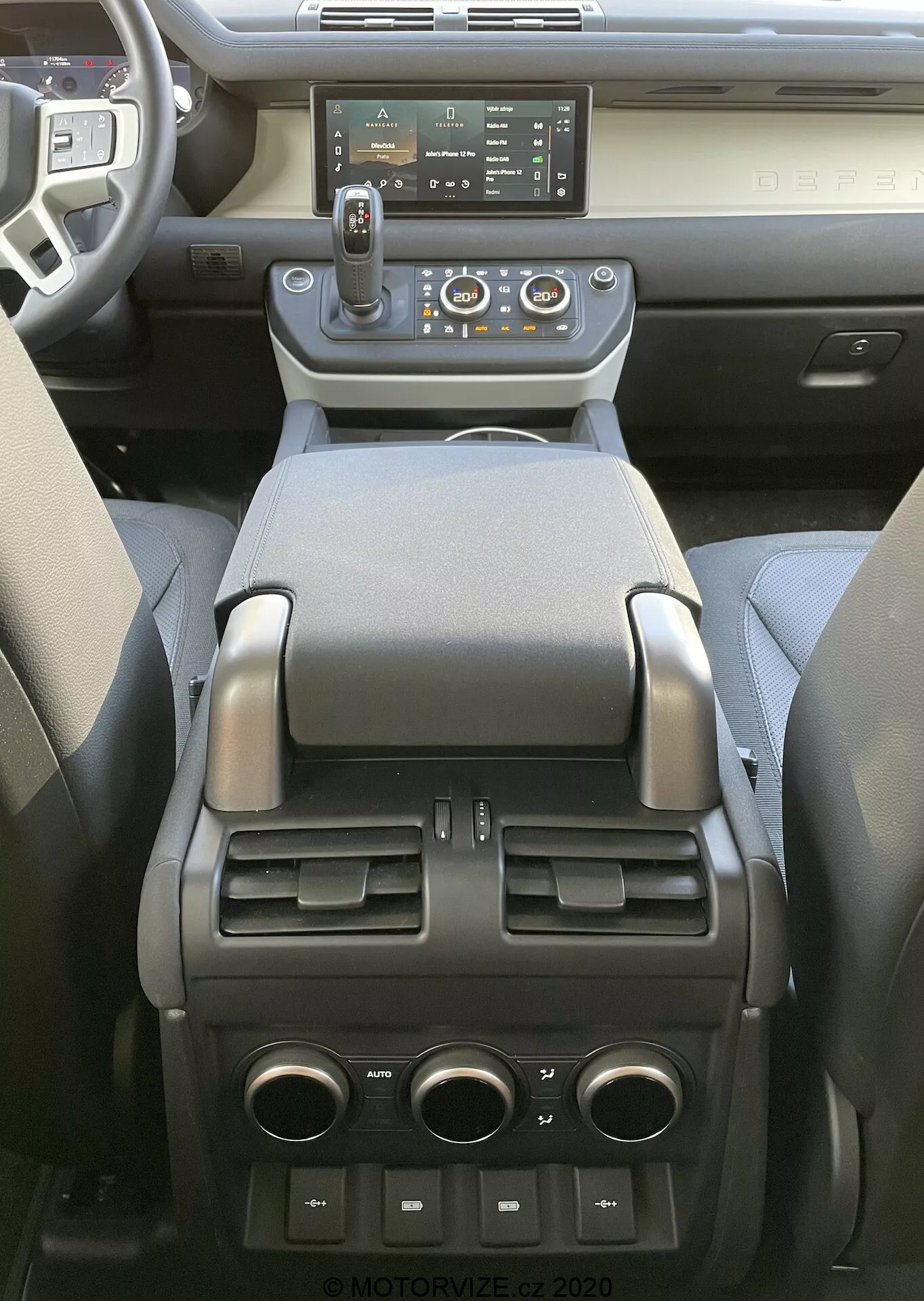 Vista interna della Land Rover Defender 2020 focalizzata sulla console centrale e la parte posteriore dell'abitacolo anteriore, mostrando un moderno sistema di infotainment touchscreen, prese d'aria centrali con pulsanti di controllo del clima, leva del cambio automatico con opzione di selezione manuale, vari pulsanti di controllo, portabicchieri, bracciolo morbido, vano portaoggetti, prese d'aria per il controllo del clima per i passeggeri posteriori e porte USB e una presa da 12V nella parte posteriore, indicanti una combinazione di tecnologia avanzata, comfort e praticità.