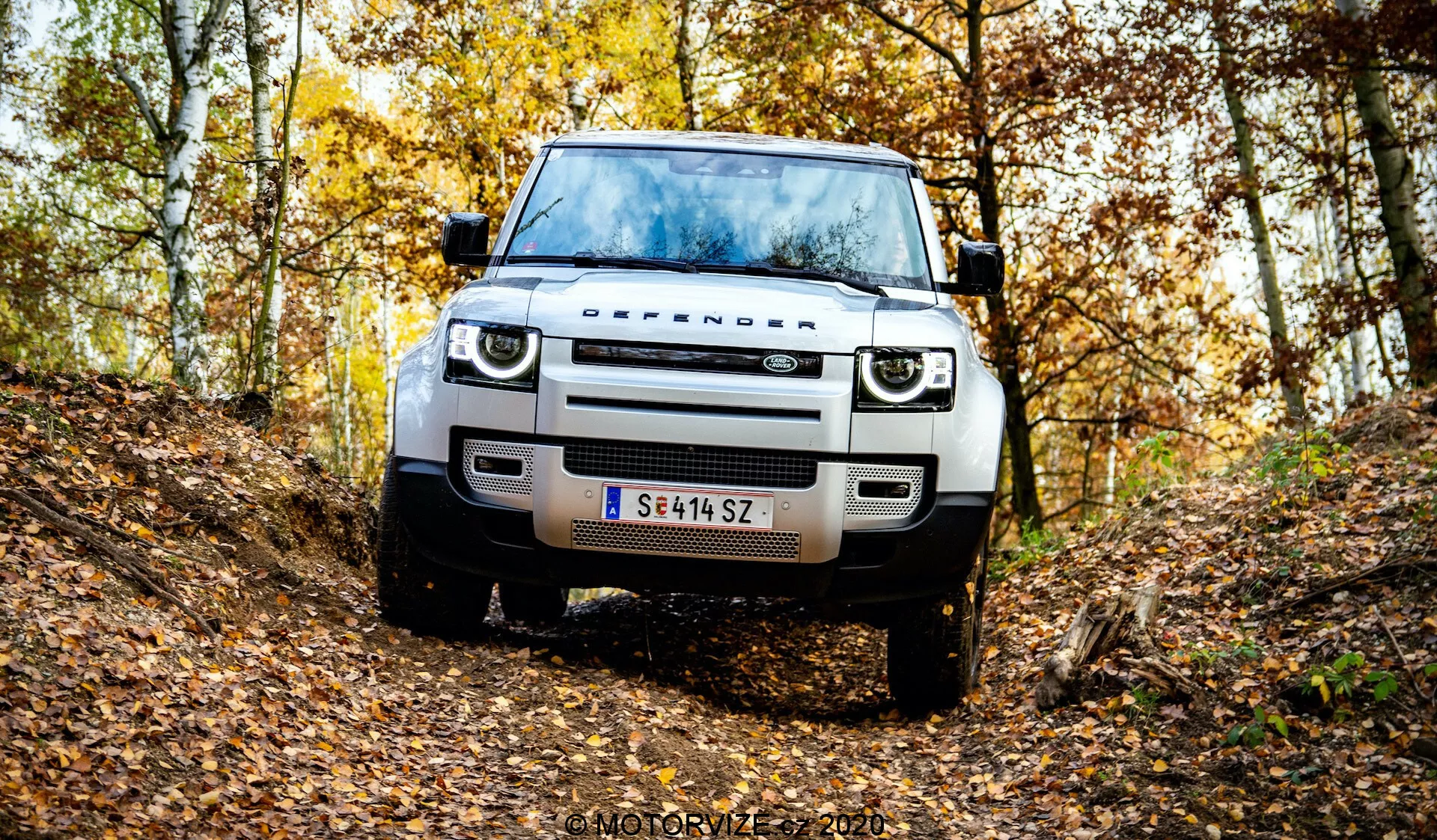 Vista frontale di una moderna Land Rover Defender in un ambiente naturale autunnale su un sentiero di terra irregolare. L'immagine cattura la griglia, i fari, il cofano, le ruote e i pneumatici del veicolo, mettendo in risalto la sua altezza da terra e le capacità fuoristrada. Caratterizzato da illuminazione LED moderna, design della griglia semplice, sensori sul paraurti anteriore o assistente al parcheggio, piastra di protezione in acciaio e targa austriaca. Le foglie cadute e gli alberi con fogliame giallo e marrone creano un'atmosfera stagionale. Filigrana 'MOTORYZE 2021' in basso a destra.