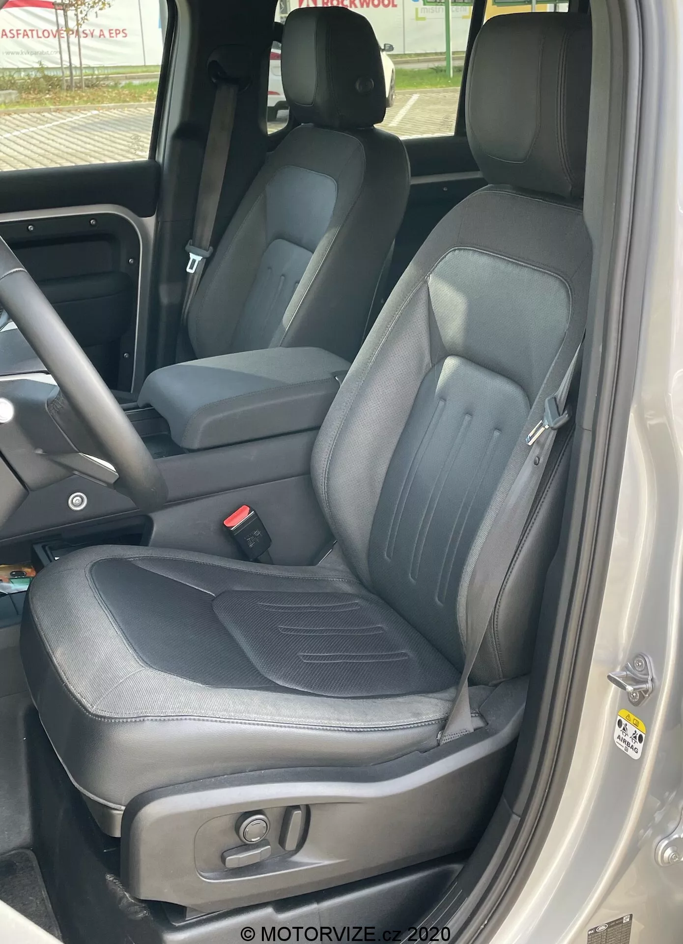 Vista ravvicinata degli interni di una Land Rover Defender 2020 focalizzata sul sedile del conducente, scattata dall'esterno dell'auto con la porta aperta mostrando tappezzeria in pelle nera con dettagli di cucitura visibili, supporti laterali, poggiatesta integrato e regolabile, controlli elettronici per la regolazione del sedile, parte della console centrale che indica un'ampia cabina anteriore e il pannello della porta con maniglia, pulsanti di controllo e altoparlanti.