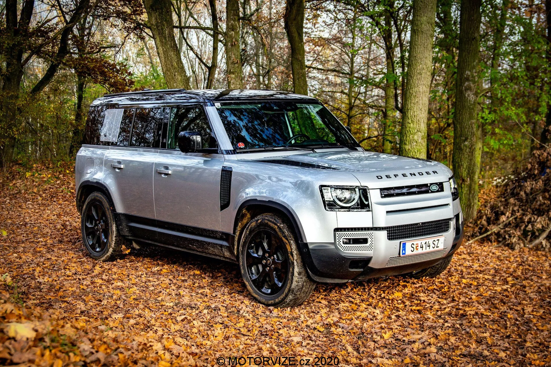 Land Rover Defender 2020 in angolazione frontale tre quarti, che mostra il suo design robusto e le caratteristiche avanzate in un ambiente naturale autunnale con foglie cadute e alberi. Il SUV ha una carrozzeria argento con dettagli neri, fari LED, tetto panoramico e cerchi in lega neri. L'immagine cattura il lato del conducente, evidenziando la griglia anteriore con il logo DEFENDER, specchietti laterali neri con indicatori e possibili piastre anti-sfregamento per fuoristrada.