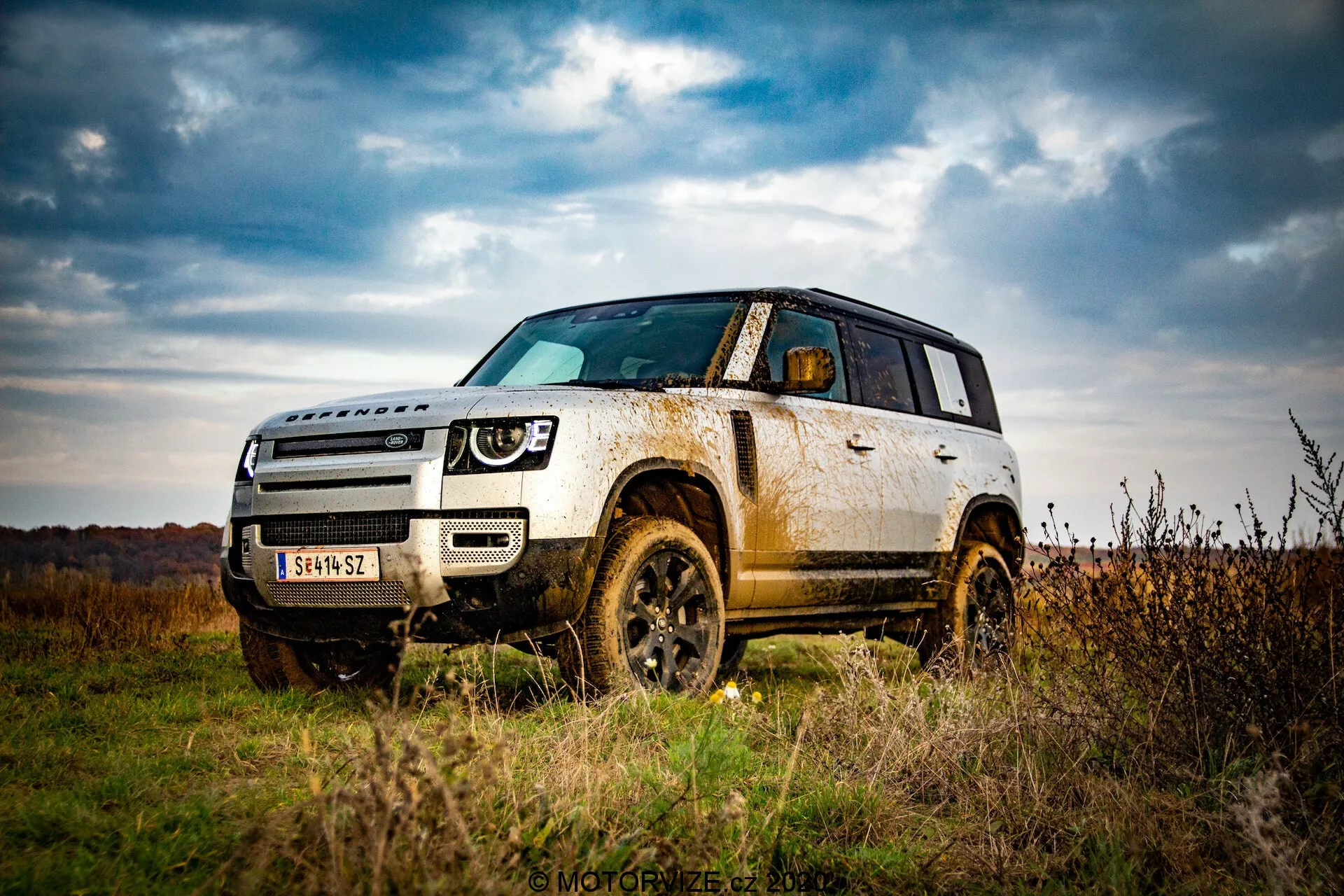Immagine di un SUV Land Rover Defender 2020 all'aperto, che evidenzia le sue capacità fuoristrada da una prospettiva a basso angolo. Enfatizza l'altezza da terra del veicolo, la robustezza con archi delle ruote, pneumatici ed elementi protettivi nella parte inferiore della carrozzeria. Cattura una vista frontale di tre quarti, mostrando l'intera parte anteriore e il lato fino alla porta posteriore, accentuando gli elementi di design come i fari, la griglia e le linee di contorno sul cofano. Il fango e la sporcizia sui pannelli laterali e sulle ruote suggeriscono un uso fuoristrada, alludendo a durabilità e capacità di tutti i terreni. I cieli nuvolosi e un paesaggio autunnale sullo sfondo aumentano la sensazione di avventura.