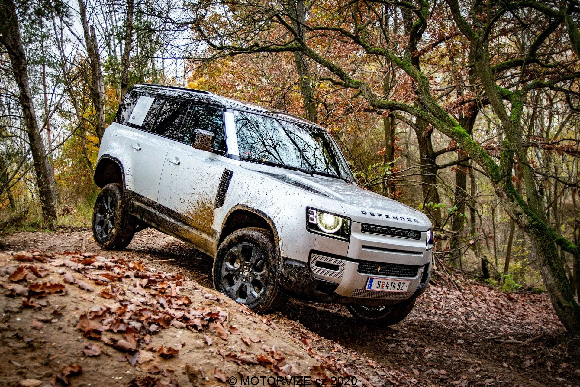 Prospettiva laterale di un SUV Land Rover Defender 2020 bianco in un ambiente off-road; design distintivo con vista della griglia frontale e del paraurti, fari LED moderni, cerchi in lega neri con pneumatici da fuoristrada, specchietti laterali dispiegati, silhouette squadrata che mostra sporco, maniglie delle porte a filo, linea del tetto piatta e alta altezza da terra. Veicolo in salita che mostra le sue capacità off-road.