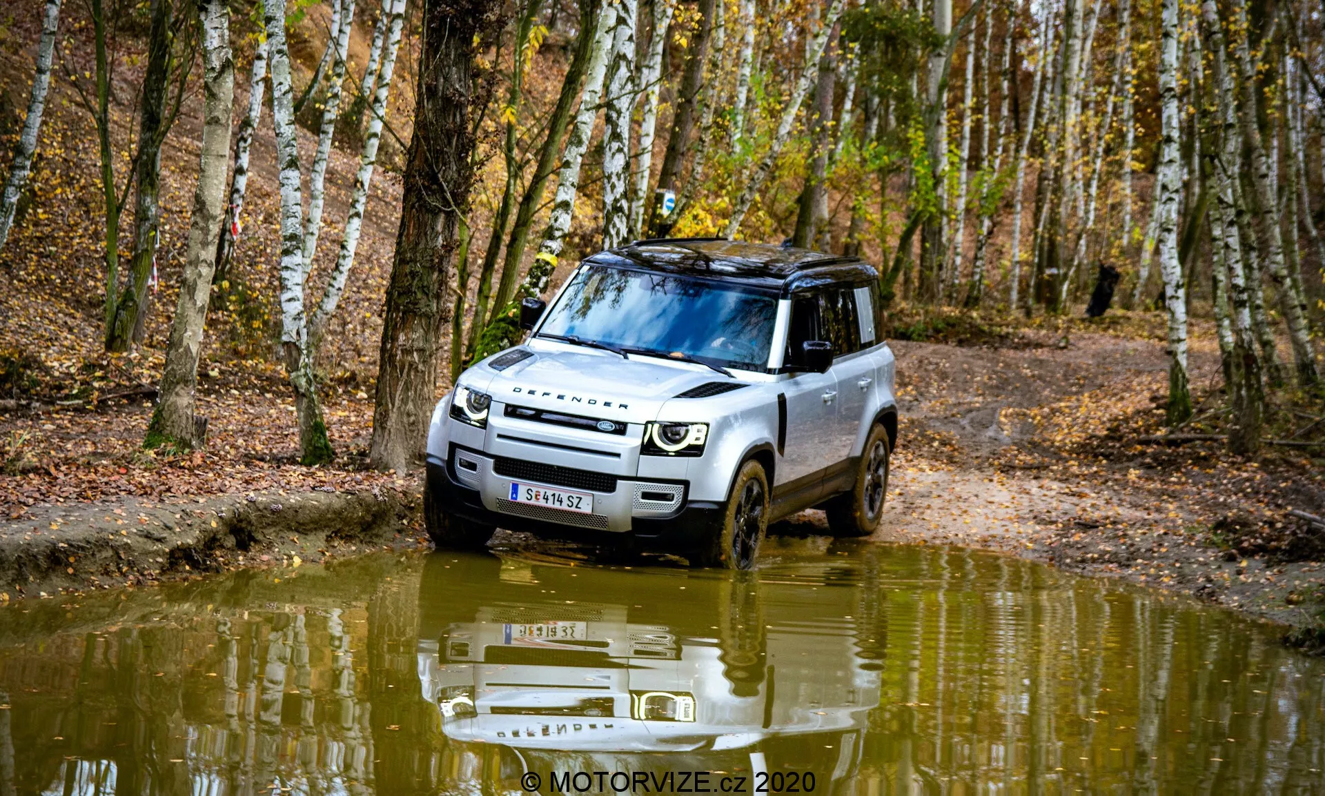 Immagine di una Land Rover Defender 2020 in un'ambientazione forestale, catturata da un'angolazione frontale a tre quarti. Il veicolo dimostra capacità fuoristrada guidando su un percorso fangoso e pieno d'acqua. Il design presenta fari moderni, una griglia prominente e una protezione sottoscocca. Barre sul tetto, pneumatici con battistrada profondo e una superficie riflettente dell'acqua aggiungono un elemento estetico. Circondato da betulle e foglie cadute, l'immagine cattura l'essenza dell'autunno.