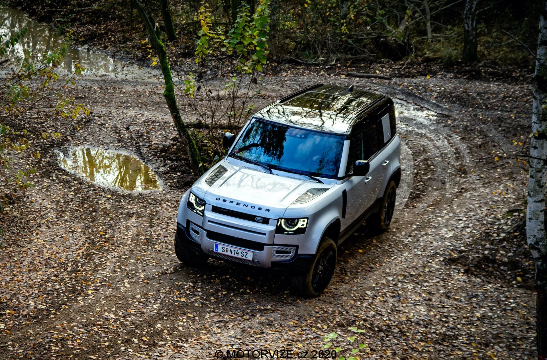 Vista dall'alto di una Land Rover Defender 2020 che naviga su un sentiero fuoristrada, con dettagli prominenti come la griglia anteriore, il cofano, il parabrezza e parte del lato del conducente, circondata da un ambiente naturale con pozzanghere d'acqua e fango, e alberi.