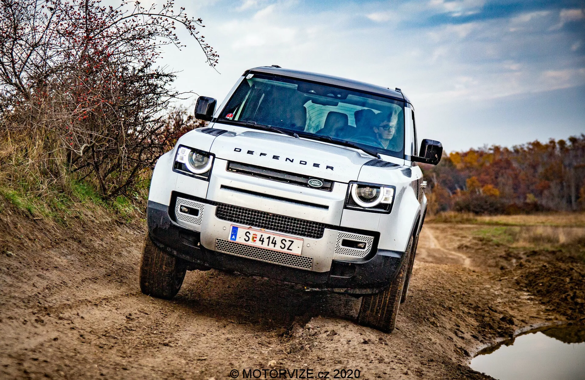 Vista tre quarti anteriore del Land Rover Defender 2020, SUV bianco e squadrato, off-road con cofano piatto, passaruota quadrati, luci diurne a LED circolari, fari adattivi, griglia a nido d'ape con logo della marca, piastra di protezione sul paraurti anteriore, assetto elevato e robusto, specchietto laterale con indicatori di direzione integrati e enfasi su griglia, fari e paraurti anteriore. Veicolo su un sentiero sterrato con sporco sull'esterno, circondato da ambiente terroso e autunnale. Foto professionale ben illuminata con sfondo sfocato e filigrana 'MOTORVIZE © 2020'.
