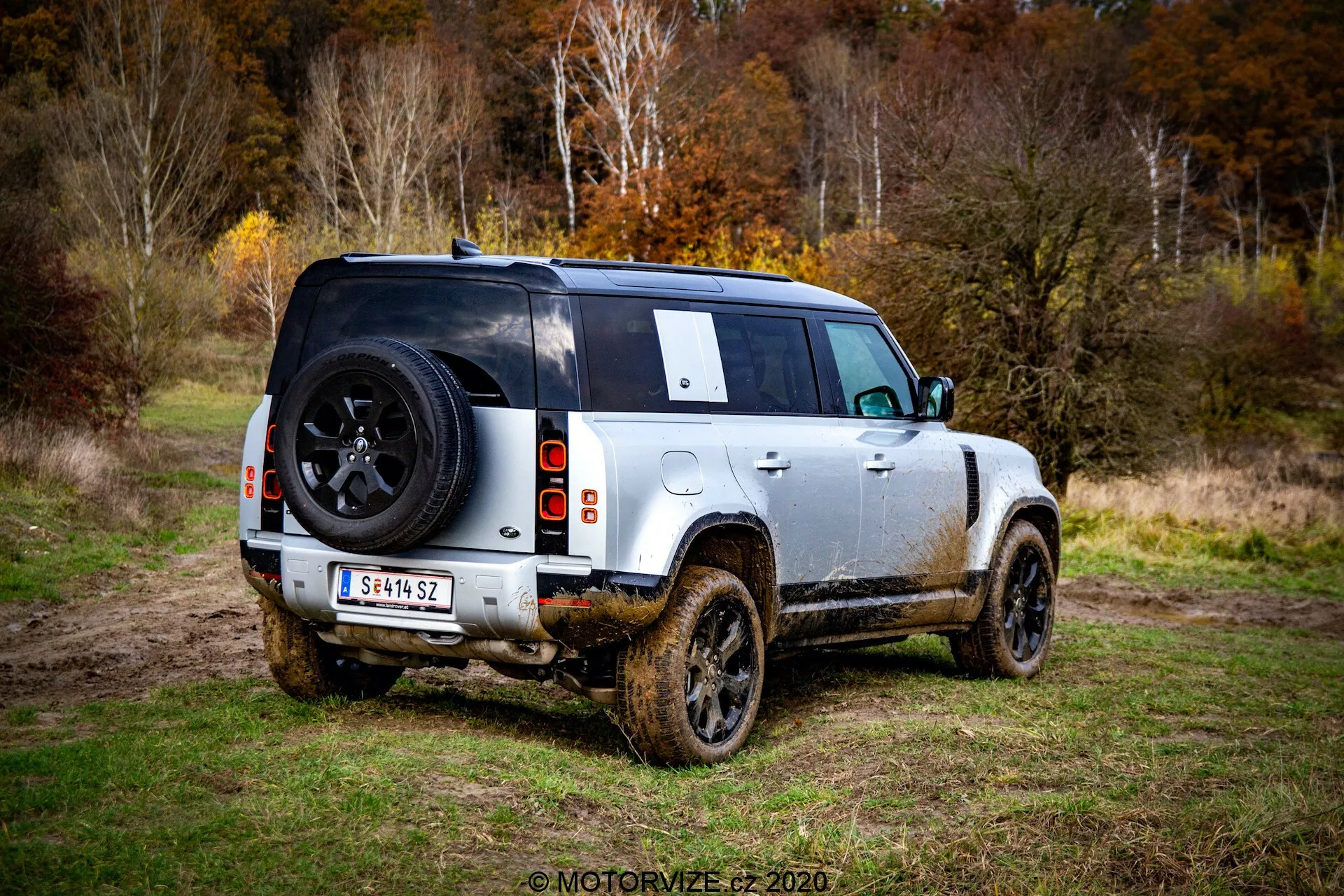 Vista posteriore tre quarti di un moderno Land Rover Defender 2020, un SUV quattro porte parcheggiato su un'area erbosa con fango visibile sui lati, indicando un recente utilizzo fuoristrada. La foto scattata dal livello del suolo mette in evidenza i profili posteriori e laterali, concentrandosi sul design posteriore inclusi una luce posteriore visibile, ruota posteriore, area di carico, e una ruota di scorta montata sulla porta posteriore con un logo del marchio. Il veicolo mostra uno schema di colori bicolore con corpo chiaro e tetto nero, cerchi in lega con robusti pneumatici per tutti i terreni, alta altezza da terra, e un finestrino posteriore parzialmente oscurato dalla ruota di scorta e un tergilunotto posteriore. I lati e i pannelli inferiori infangati suggeriscono che il SUV è stato guidato in terreno fangoso. Sullo sfondo si intravede un'area boschiva con luce soffusa, indicando un cielo coperto o condizioni di scarsa luce.