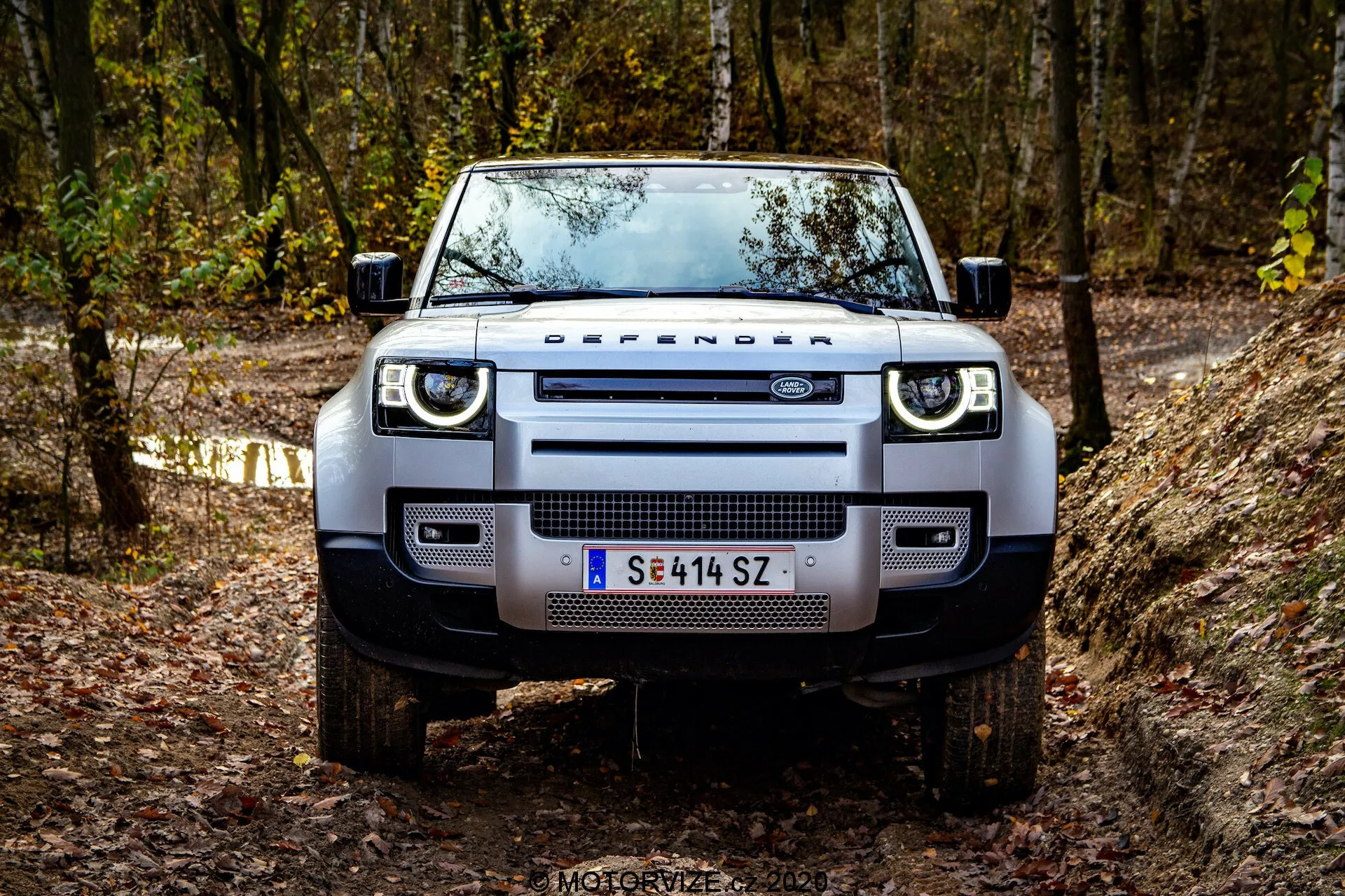 Vista frontale di una Land Rover Defender 2020 su un sentiero fuoristrada, che mostra la sua presenza robusta e dominante. I dettagli visibili includono la distintiva griglia con la scritta 'DEFENDER', fari rettangolari moderni con luci diurne a LED, ampio paraurti anteriore con luci fendinebbia integrate, robuste piastre di protezione, cofano piatto con bordi pronunciati, pneumatici da fuoristrada montati su ruote in lega e specchietti laterali funzionali con indicatori di direzione integrati. Il veicolo è situato in un'area forestale con fogliame autunnale, evidenziando il suo spirito avventuroso e fuoristrada.