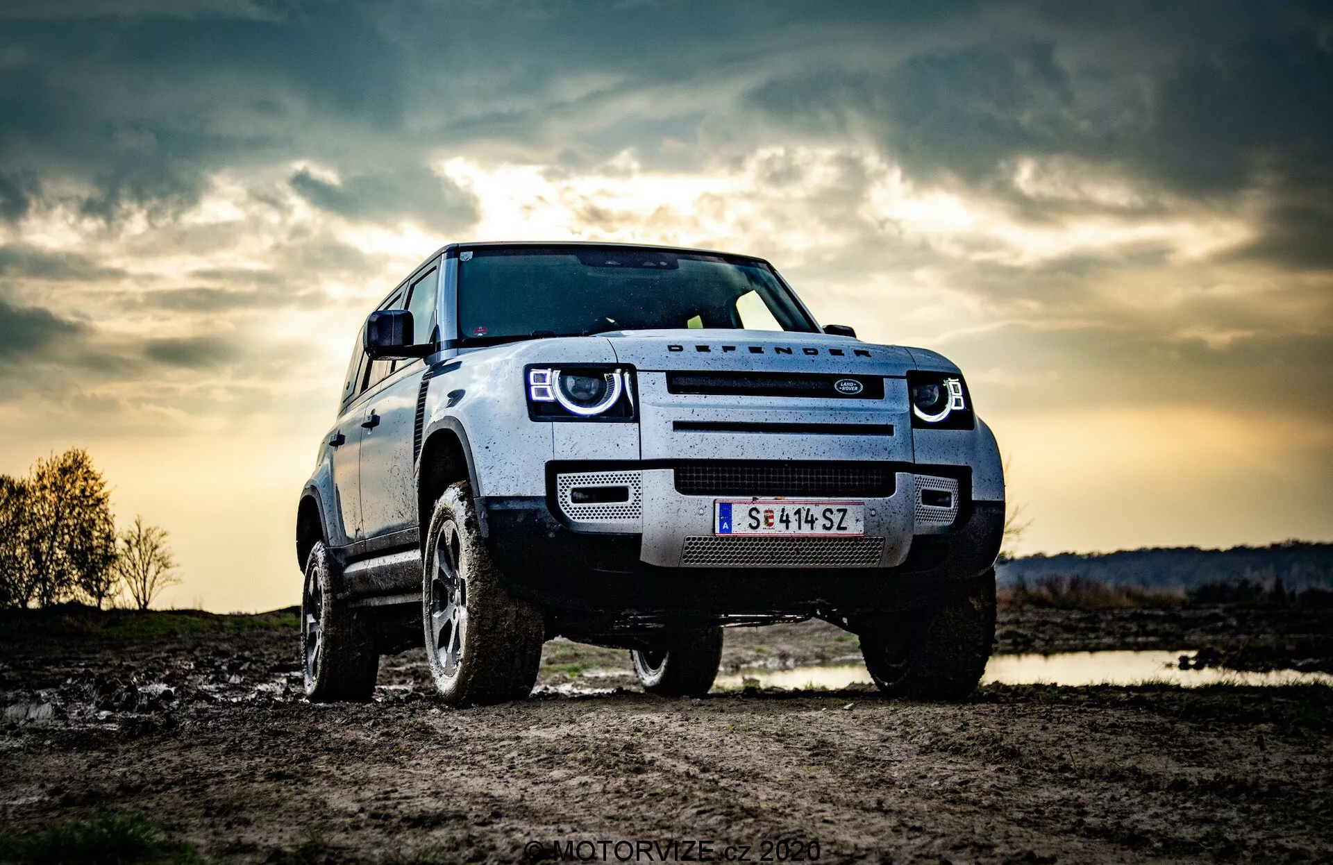 L'immagine mostra un SUV Land Rover Defender 2020 parcheggiato all'aperto, possibilmente in un contesto fuoristrada. Catturato da una prospettiva a bassa angolazione, mette in risalto la robusta statura e l'altezza del veicolo. La vista anteriore a tre quarti mostra la griglia frontale, i fari, il cofano e il parafango e la ruota del lato guida. Sono visibili le distintive luci diurne a LED, suggerendo caratteristiche di illuminazione moderne. La griglia frontale audace probabilmente riflette l'identità del produttore e una telecamera o sensore frontale sulla griglia indica sistemi avanzati di assistenza alla guida. Lo sfondo drammatico del cielo, possibilmente con illuminazione serale o crepuscolare, conferisce un senso di avventura, mentre le robuste ruote e l'elevata altezza dal suolo enfatizzano la capacità fuoristrada.