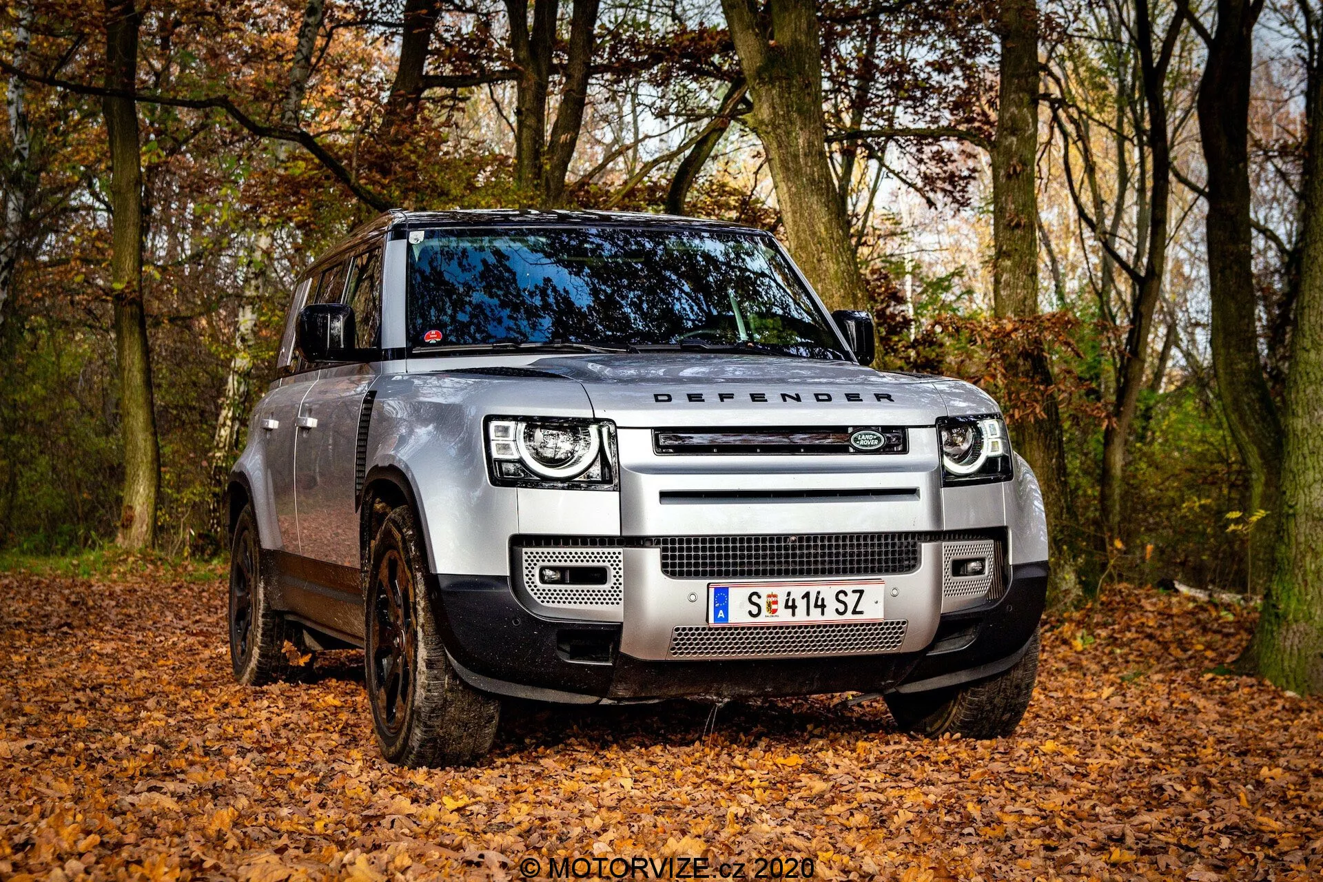 Vista angolare frontale di una moderna Land Rover Defender 2020 in un ambiente fuoristrada con foglie cadute, catturata da una prospettiva a bassa angolazione. L'immagine mette in evidenza la griglia frontale con la scritta 'DEFENDER' sul cofano, i fari a LED con luci diurne quadrate e i sensori moderni integrati nel paraurti anteriore. Le ruote coperte di fango e le sospensioni pneumatiche regolabili indicano la sua capacità fuoristrada. Sono visibili il lato del conducente, la porta anteriore e parte della porta posteriore, mostrando il design robusto del veicolo con ruote parzialmente girate.