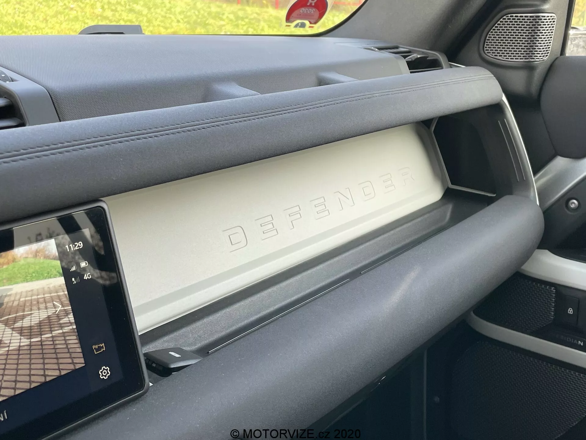 Interno del Land Rover Defender 2020 che mostra il lato passeggero del cruscotto dall’angolo del sedile del conducente. L'immagine cattura il vano portaoggetti chiuso con il rilievo 'DEFENDER', che esemplifica il design lussuoso e robusto del veicolo. A destra, un moderno display touchscreen di infotainment mostra i controlli del lettore multimediale e una mappa di navigazione, indicando caratteristiche tecnologiche avanzate. I materiali del cruscotto dimostrano una combinazione di texture premium e pratiche, e la griglia dell'altoparlante è visibile a sinistra, integrandosi con il design robusto degli interni.