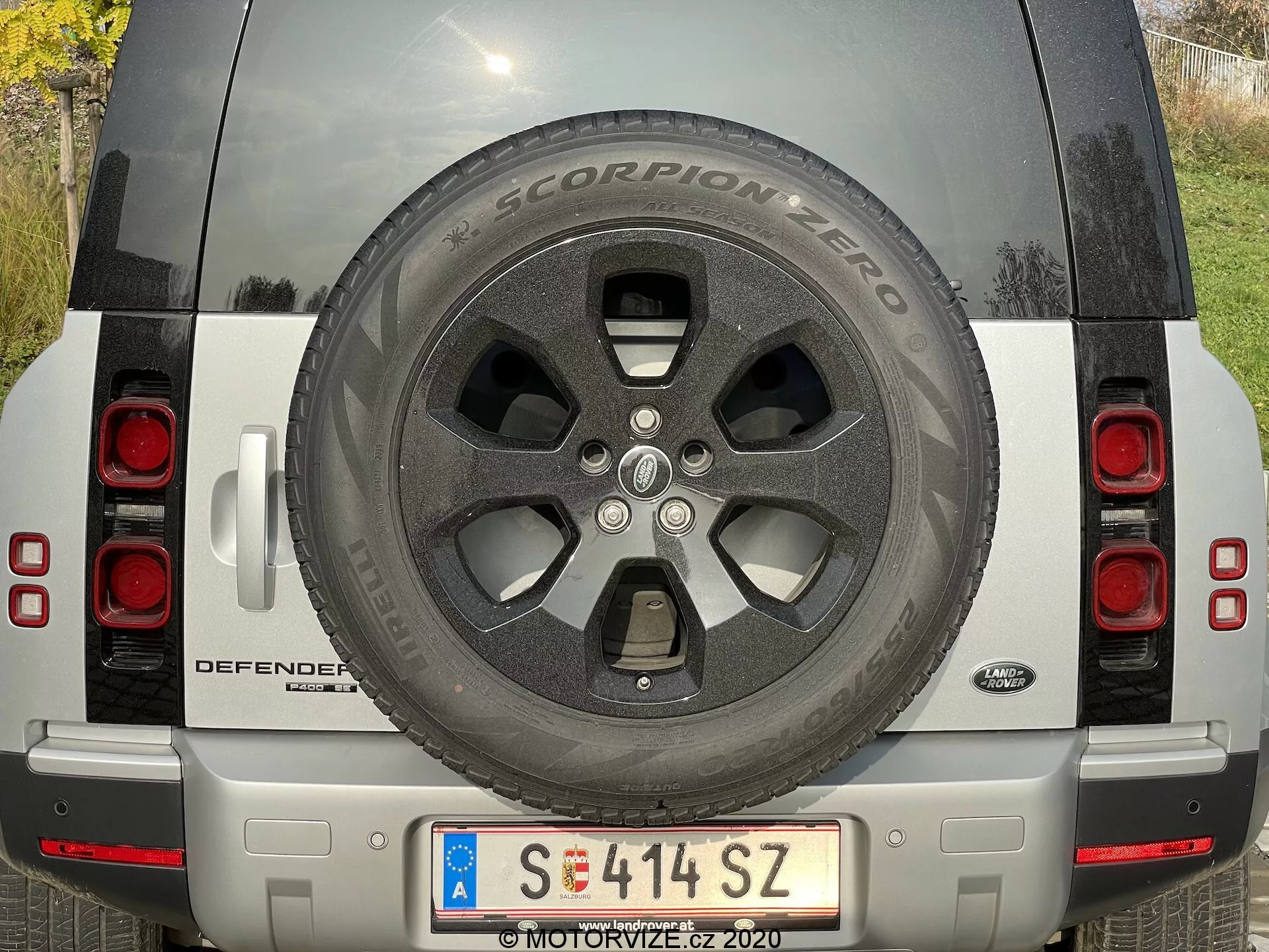 Vista posteriore di una Land Rover Defender 2020 argento con ruota di scorta visibile, luci posteriori, targa, nome del modello DEFENDER e badge Land Rover, paraurti argento con sensori e telecamera e riflettori. Il SUV è parcheggiato alla luce naturale del giorno, evidenziando le condizioni pulite e ben mantenute del veicolo.