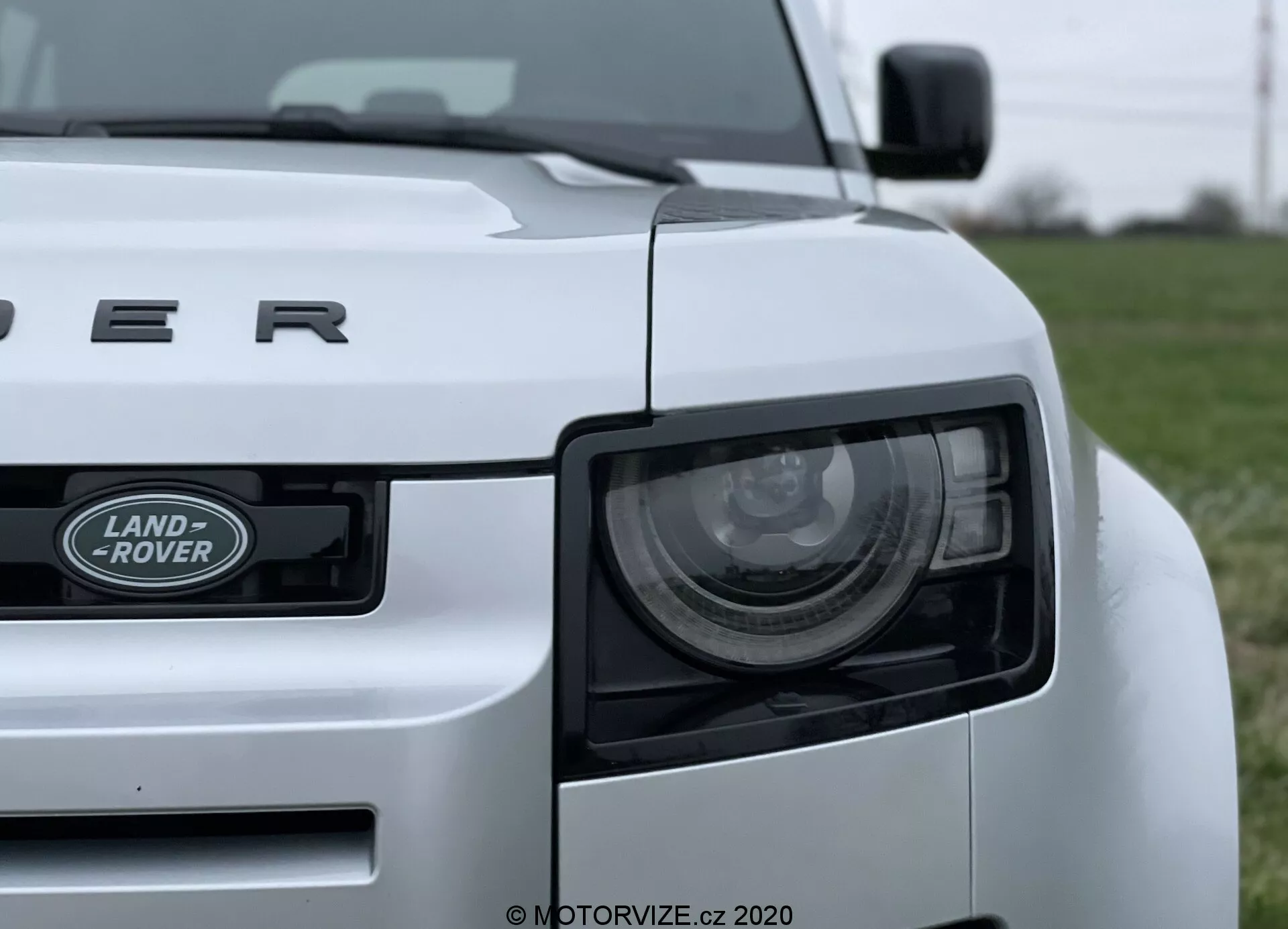 Immagine ravvicinata di una Land Rover Defender 2020 bianca, presa da un angolo basso, che mostra il quarto anteriore sinistro del veicolo. Le caratteristiche principali includono il faro, la griglia con l'emblema 'LAND ROVER' e il cofano con la scritta 'DEFENDER'. Il faro presenta elementi LED moderni, e il design robusto della vettura è evidenziato da linee e curve visibili. L'auto è posizionata su un campo erboso, con lo sfondo sfocato per enfatizzare il veicolo.