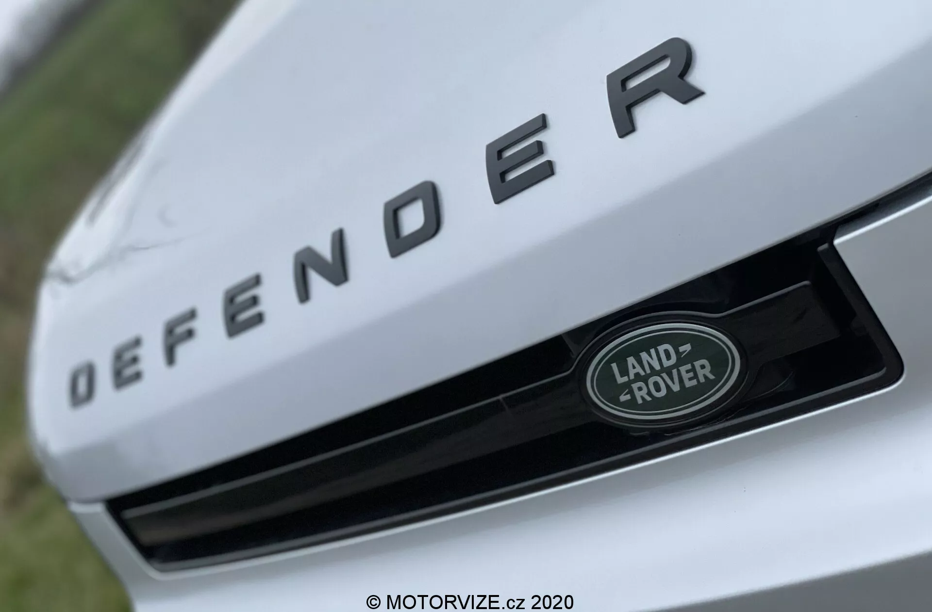 Vista ravvicinata della sezione anteriore di un Land Rover Defender 2020, con un focus prominente sulla targhetta con il nome 'DEFENDER' in lettere argentate in rilievo sul bordo anteriore del cofano. Sotto la targhetta, il badge Land Rover in argento e verde con bordo bianco è posizionato centralmente sulla griglia. L'immagine è scattata da un angolo basso e ascendente con una ridotta profondità di campo, mettendo in evidenza gli elementi di branding mentre lo sfondo è sfocato.