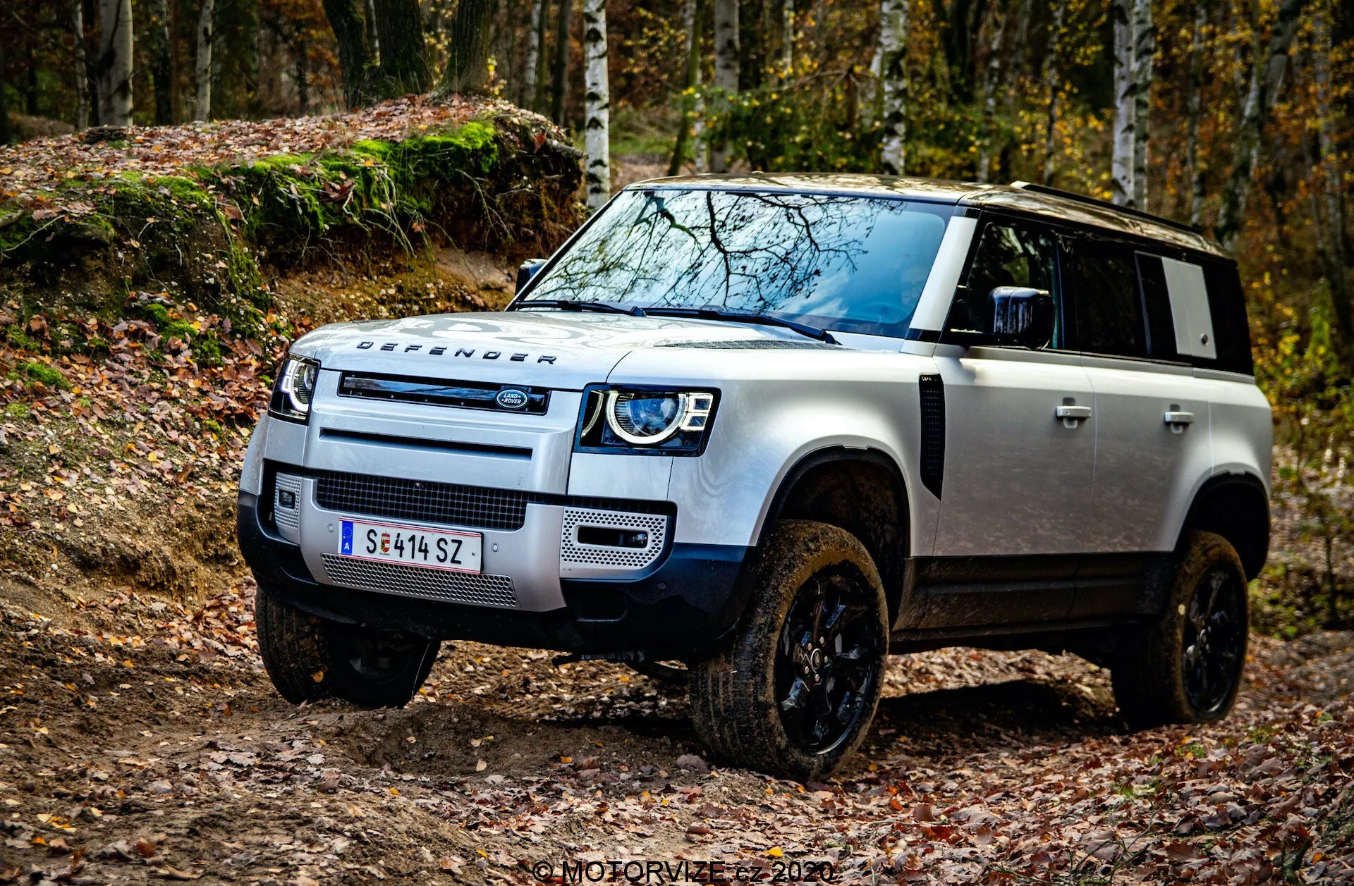 Scatto da un'angolazione bassa di un Land Rover Defender 2020 ripreso dal quarto anteriore sinistro, evidenziando le caratteristiche fuoristrada robuste come la griglia frontale distintiva di Land Rover, fari a LED con luci diurne, piastra di protezione sul paraurti anteriore, marchio DEFENDER prominente sul cofano, cofano nero a contrasto su carrozzeria bianca, prese d'aria rialzate per traversate in acque profonde, grandi passaruota con ruote fuoristrada nere, indicatori di direzione sugli specchietti laterali e tecnologia avanzata di assistenza alla guida vicino al paraurti anteriore. L'immagine è ambientata in un contesto fuoristrada con foglie cadute, enfatizzando l'estetica avventurosa del veicolo. Filigrana 'MOTORVIZA 2021'.