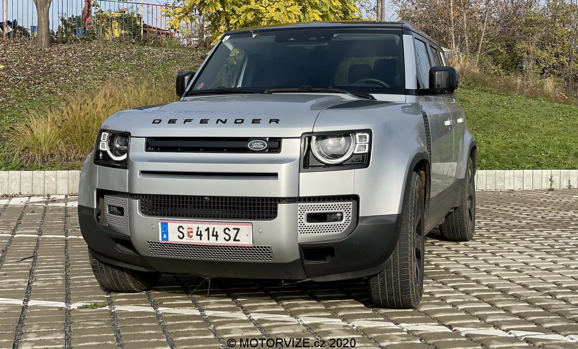 Vista frontale di una Land Rover Defender modello 2020, con fari distintivi a LED a due livelli, griglia prominente con logo Land Rover, paraurti geometrico con piastra di protezione argentata e altre caratteristiche robuste che suggeriscono capacità fuoristrada. Visibili anche il cofano con la targa 'Defender', le prese d'aria e viste parziali delle ruote a cinque razze. Il veicolo è di colore neutro chiaro, probabilmente argento o grigio.