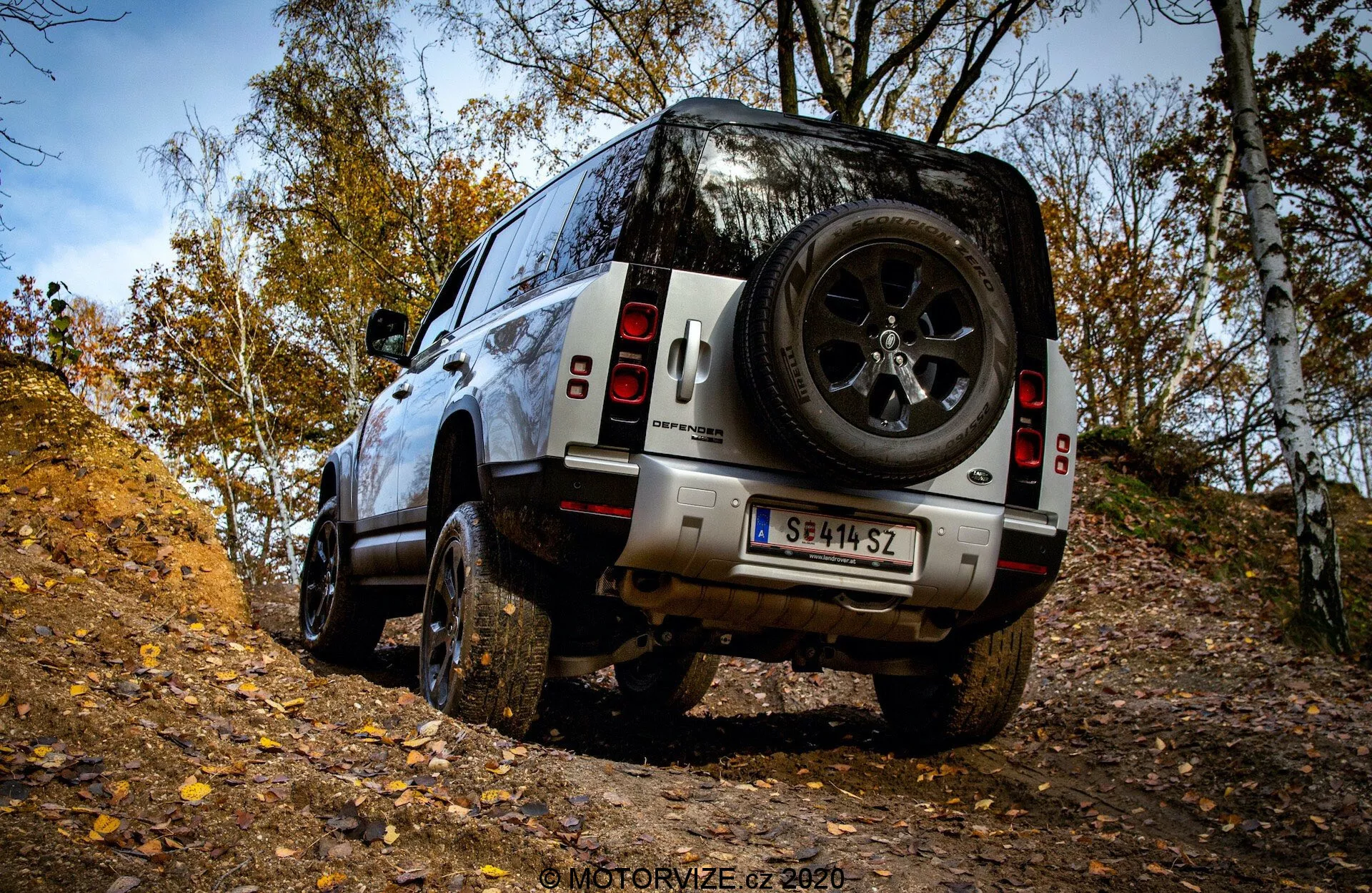 Vista posteriore tre-quarti del SUV Land Rover Defender 2020 su terreno fuoristrada, che mostra pneumatici robusti, ampia altezza da terra e ruota di scorta posteriore in un ambiente forestale autunnale.