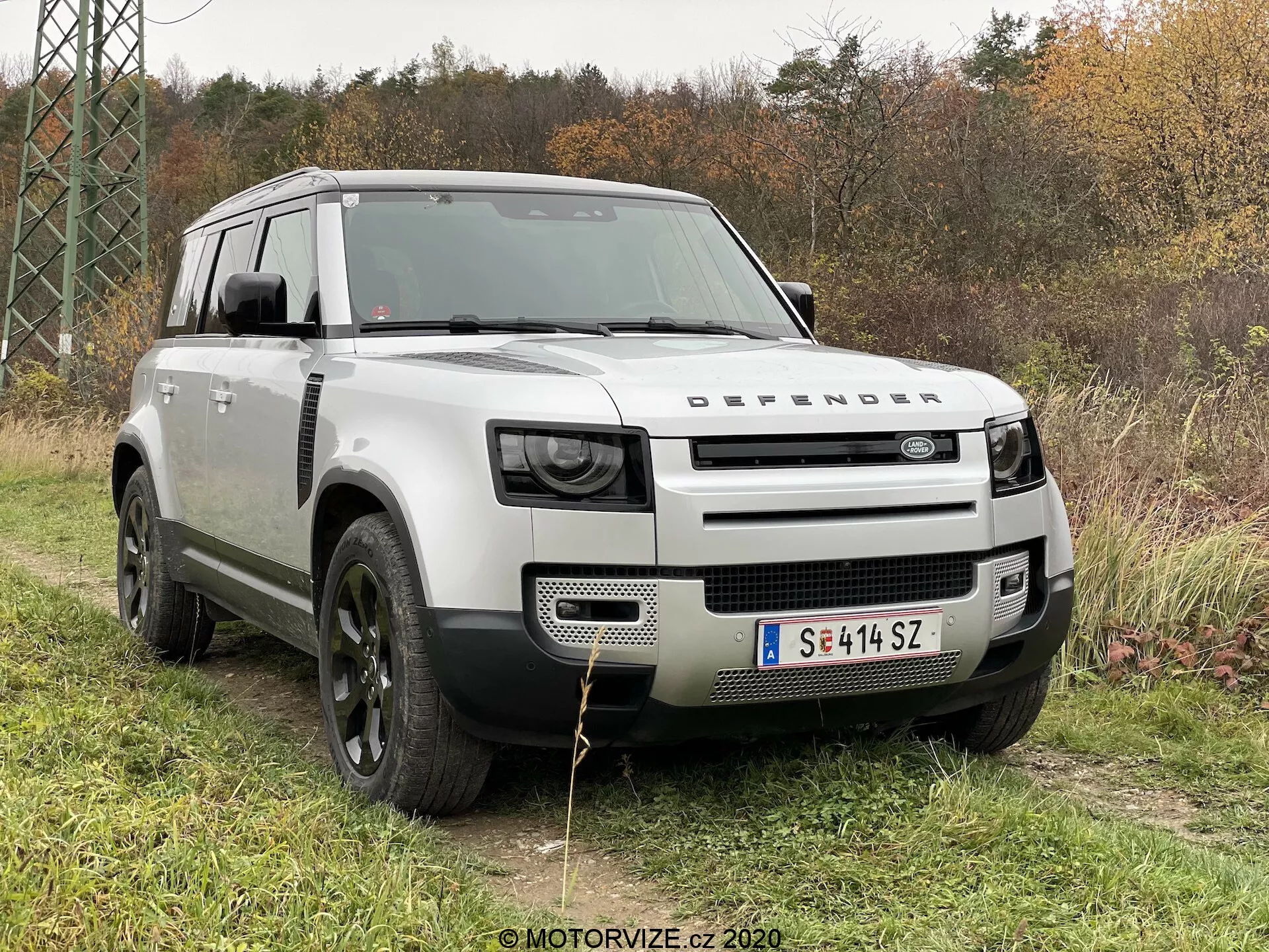 Questa immagine presenta una Land Rover Defender 2020 in un ambiente all'aperto su un prato con fogliame e alberi sullo sfondo. Catturata da un angolo anteriore di tre quarti, la foto mette in risalto la griglia prominente con il marchio 'DEFENDER', i fari LED moderni, il robusto cofano con design a conchiglia, le grandi ruote in lega nera con pneumatici fuoristrada e una carrozzeria color argento chiaro che contrasta con i passaruota e il tetto neri. Elementi di design come la piastra antincastro anteriore, le finestre di aspirazione 'alpine' e la struttura squadrata enfatizzano le sue capacità fuoristrada e utilitarie.
