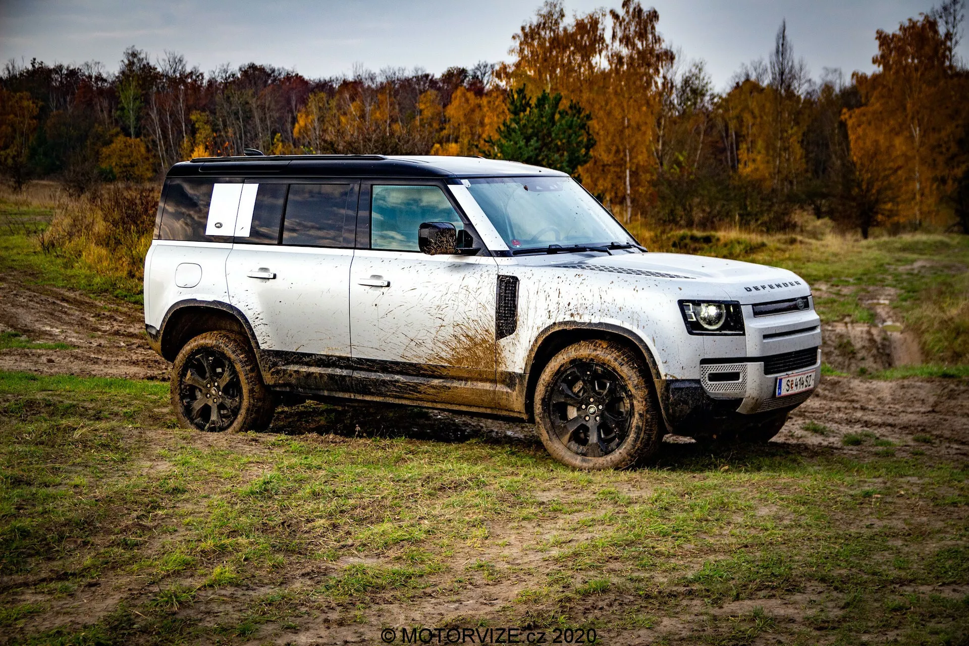 Profilo laterale di un SUV fuoristrada Land Rover Defender 2020 mostrato da un angolo di tre quarti, con la carrozzeria bianca con tetto e ruote nere, coperto di fango. Veicolo posizionato diagonalmente da sinistra in basso a destra in alto contro un terreno naturale fangoso sotto un cielo coperto con alberi autunnali sullo sfondo.