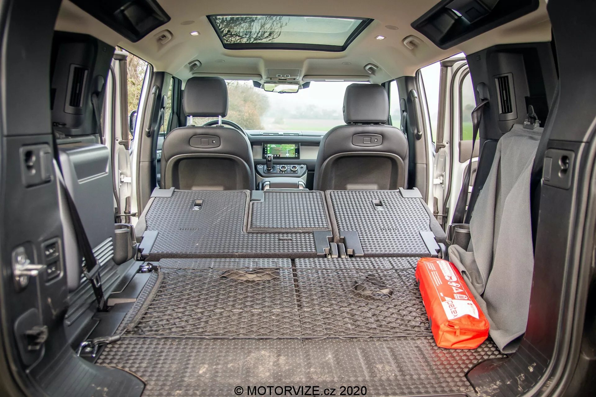 Vista interna dello spazio di carico del Land Rover Defender 2020 presa da un angolo rivolto verso il retro, che mostra il tappetino protettivo con motivi a piastra diamantata che copre l'intero pavimento del vano di carico. I sedili posteriori sono ripiegati in piano, rivelando un ampio spazio di carico. Le pareti laterali presentano accessori come altoparlanti e portabicchieri, suggerendo un design equilibrato tra funzionalità e comfort. Il cruscotto anteriore a distanza è parzialmente visibile, con il sistema di infotainment e i controlli del clima. Un kit di emergenza rosso è posizionato in modo prominente sul tappetino del vano di carico, suggerendo l'uso del veicolo per famiglie o avventure. La porta posteriore aperta permette alla luce naturale di entrare nell'area di carico.