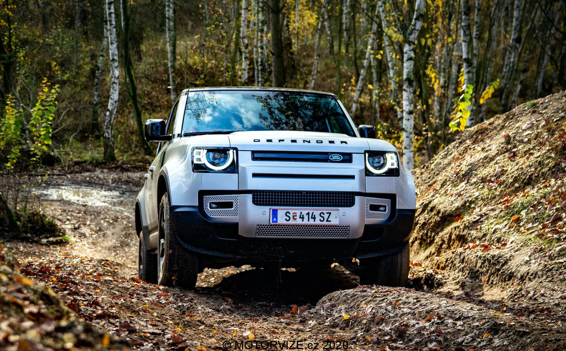 Vista frontale di un Land Rover Defender 2020, catturata da un angolo basso che enfatizza la griglia, i fari e il paraurti anteriore. Il SUV è posizionato su una strada sterrata con una foresta sullo sfondo, evidenziando la sua prontezza per il fuoristrada. Gli elementi visibili includono il design prominente 'DEFENDER', fari a LED, parafanghi larghi e pneumatici robusti.