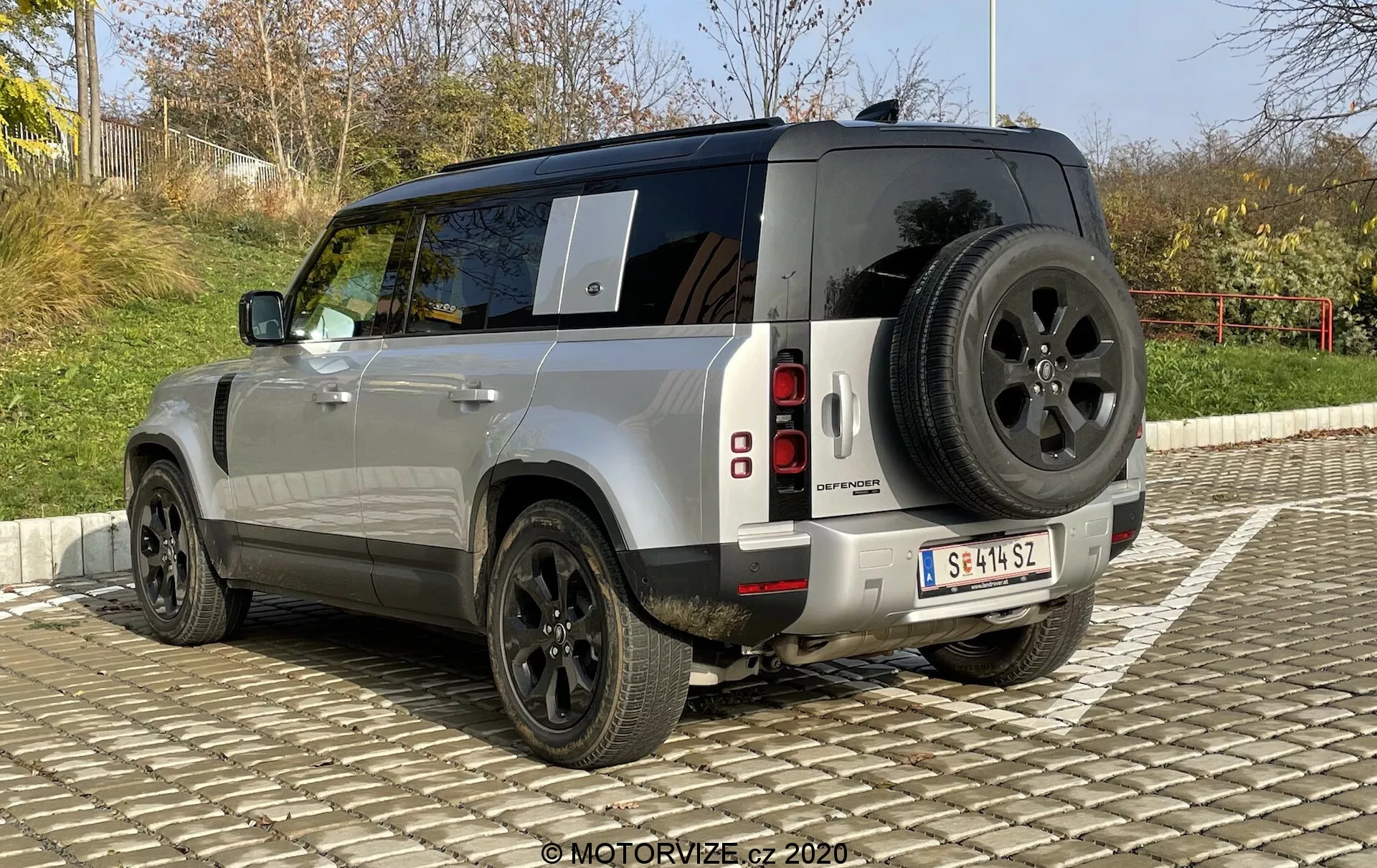 L'immagine mostra la vista di tre quarti posteriore di un SUV Land Rover Defender 2020. Questo angolo permette una visione chiara del design squadrato e robusto del veicolo, della vernice metallica argento e delle luci posteriori a LED segmentate verticalmente. I dettagli visibili includono una ruota di scorta montata sul portellone posteriore, un robusto paraurti posteriore con sensori di parcheggio, grandi cerchi in lega neri con spessi fianchi degli pneumatici per un uso fuoristrada e vetri oscurati sui finestrini posteriori. Si nota inoltre un'antenna sul tetto, che suggerisce la presenza di sistemi di comunicazione avanzati. L'auto è parcheggiata su un'area pavimentata a incastro sotto un cielo limpido.