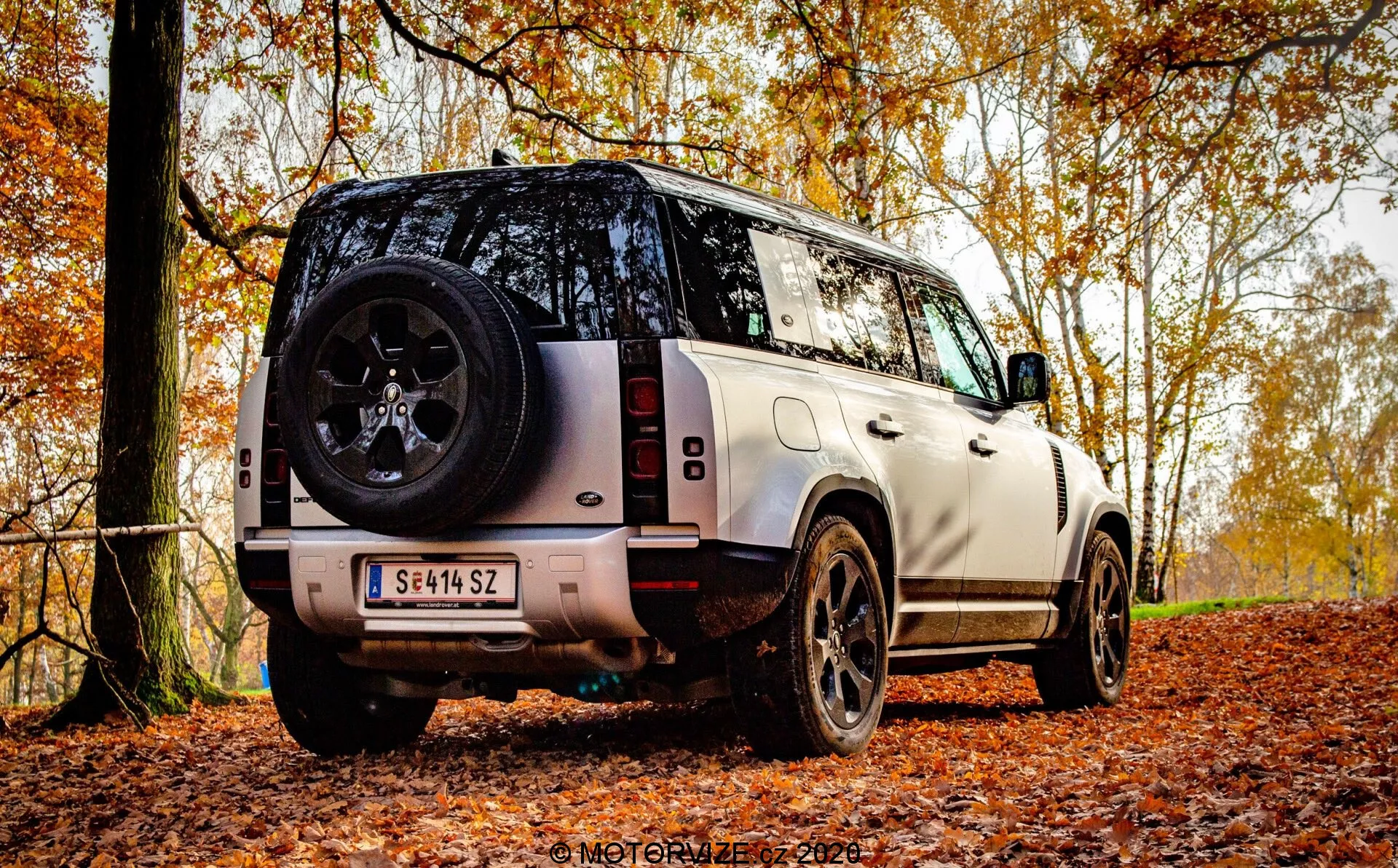 Vista posteriore a tre quarti di un SUV moderno parcheggiato su un sentiero forestale autunnale coperto di foglie, evidenziando l'altezza del veicolo e la distanza dal suolo per le capacità fuoristrada. L'inquadratura mostra il posteriore comprendente il portellone, parte del tetto, il lunotto e un profilo laterale. Le caratteristiche visibili sono il design verticale delle luci posteriori, robusti cerchi in lega neri con pneumatici a battistrada spesso, ruota di scorta sul portellone, paraurti posteriore con sensori di parcheggio, verniciatura bicolore con tetto a contrasto, tergicristallo del lunotto, telecamera posteriore sotto la targa e pedane laterali per accedere all'abitacolo. La carrozzeria squadrata e robusta trasmette lusso con una forte linea caratteristica lungo le portiere laterali. La luce naturale e diffusa suggerisce una giornata nuvolosa o un filtro della volta arborea, migliorando i temi stagionali e all'aperto.