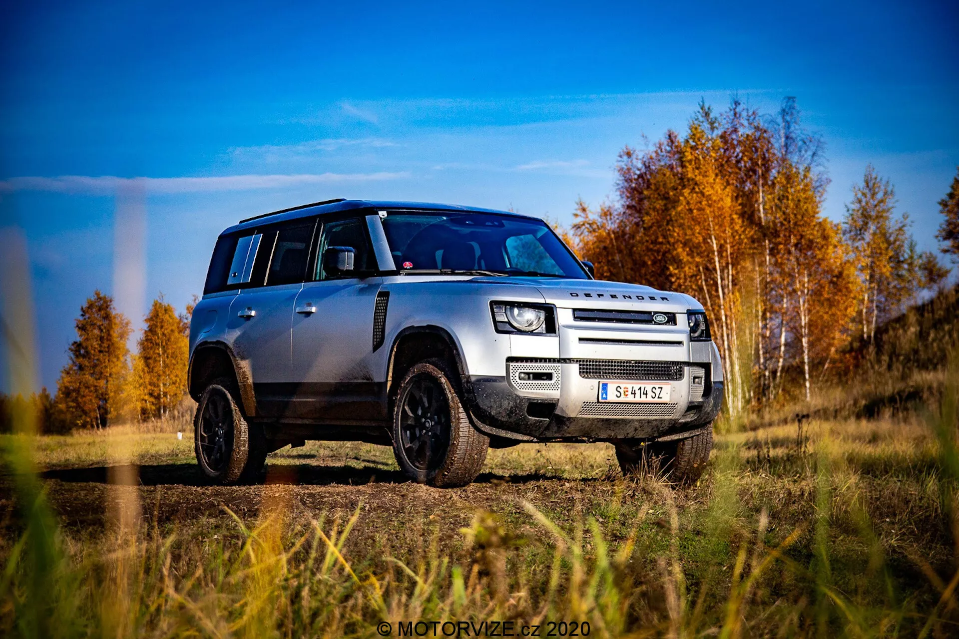 Immagine di una Land Rover Defender argentata del 2020 da una prospettiva a livello del suolo, catturata in una vista frontale obliqua di tre quarti. Il veicolo mostra chiaramente la sua griglia, il cofano, il paraurti anteriore e il design delle ruote, leggermente orientato verso il lato del conducente. Il Defender è posizionato su un terreno all'aperto con uno sfondo di alberi autunnali con foglie rosse e arancioni sotto un cielo blu parzialmente nuvoloso. Le caratteristiche di design notevoli includono la sua caratteristica forma squadrata, fari a LED moderni, fendinebbia, griglia a due barre con marchio LAND ROVER, piastra di protezione anteriore, cerchi in lega neri con pneumatici fuoristrada e un tetto nero a contrasto con calotte degli specchietti nere coordinate. La foto è estremamente nitida e dinamica, enfatizzando la robustezza e l'estetica moderna del veicolo.