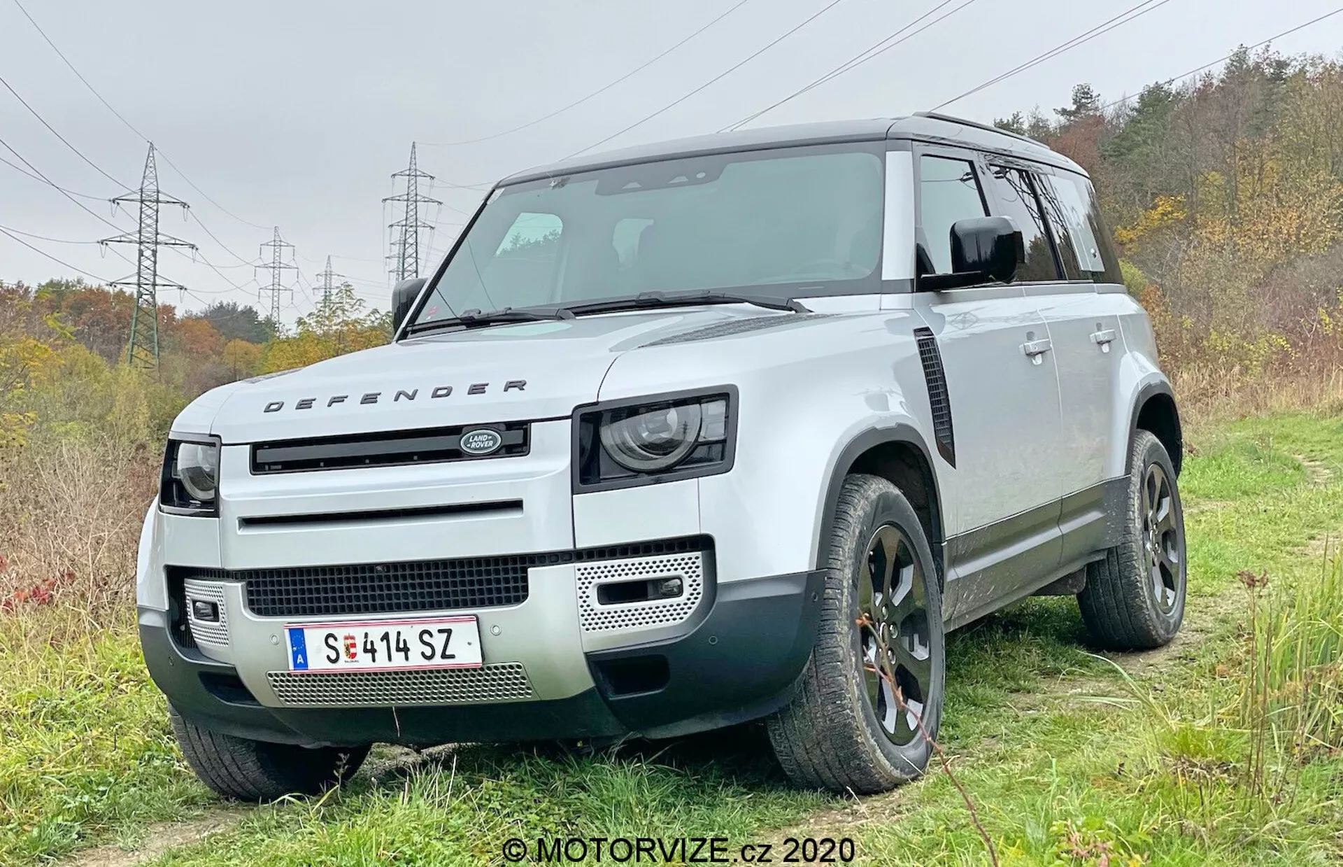 Questa fotografia cattura un SUV Land Rover Defender 2020 moderno da un'angolazione frontale di tre quarti, mostrando il lato del conducente. Il veicolo è leggermente fuoristrada in una zona erbosa con un accenno di pista sterrata, evidenziando le sue capacità fuoristrada. Il design squadrato distintivo presenta un'elevata altezza da terra, uno schema di colori bicolore con carrozzeria bianca e tetto nero a contrasto. La fascia anteriore mostra gruppi ottici a LED ispirati al retrò, un cofano con il badge Defender e una griglia anteriore minimalista, sottolineando la sua reputazione robusta e all-terrain. Le caratteristiche tecniche visibili includono sensori di parcheggio anteriori integrati nel paraurti, suggerendo sistemi avanzati di assistenza alla guida. Gli archi delle ruote robusti ospitano grandi pneumatici fuoristrada, e elementi di design come cerchi in lega e design unico della griglia sono messi in risalto.