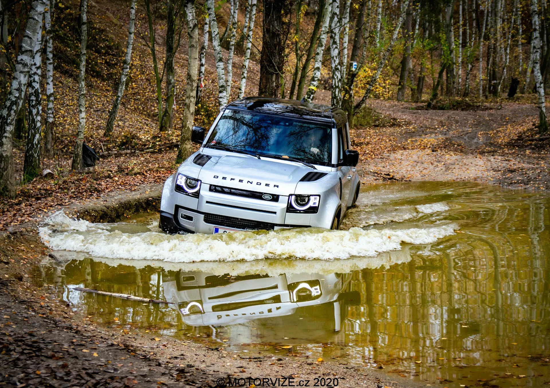 Scatto in basso angolo di una Land Rover Defender 2020 che attraversa un corpo d'acqua, evidenziando il lato anteriore destro con griglia visibile, fari a LED, cofano, parabrezza, tetto, ruota anteriore e porta del conducente. La scena dinamica cattura lo spostamento dell'acqua sottolineando le sue capacità fuoristrada, alta capacità di guado, piastra protettiva e lussuoso tetto panoramico. Il marchio Land Rover è visibile sul cofano, evidenziando il design robusto e versatile.