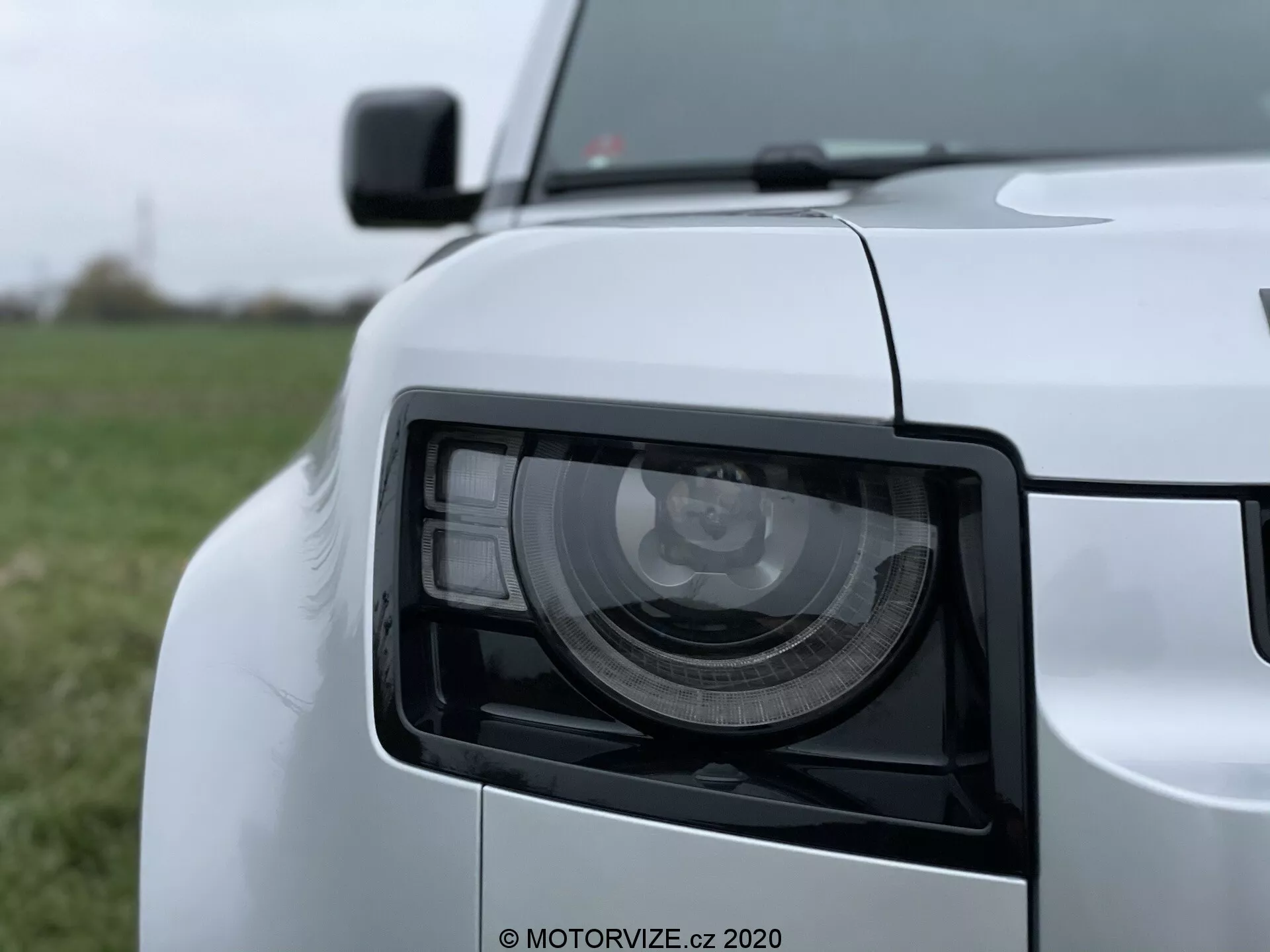 Vista ravvicinata del gruppo ottico anteriore sinistro della Land Rover Defender 2020 con un design angolare moderno, ripresa a un angolo leggero con una prospettiva da bassa a media altezza. L'immagine mostra i dettagliati moduli LED/HID, la finitura con vernice bianca lucida e parte della griglia anteriore sfocata, il bordo del cofano, la sezione del paraurti anteriore e lo specchietto laterale.