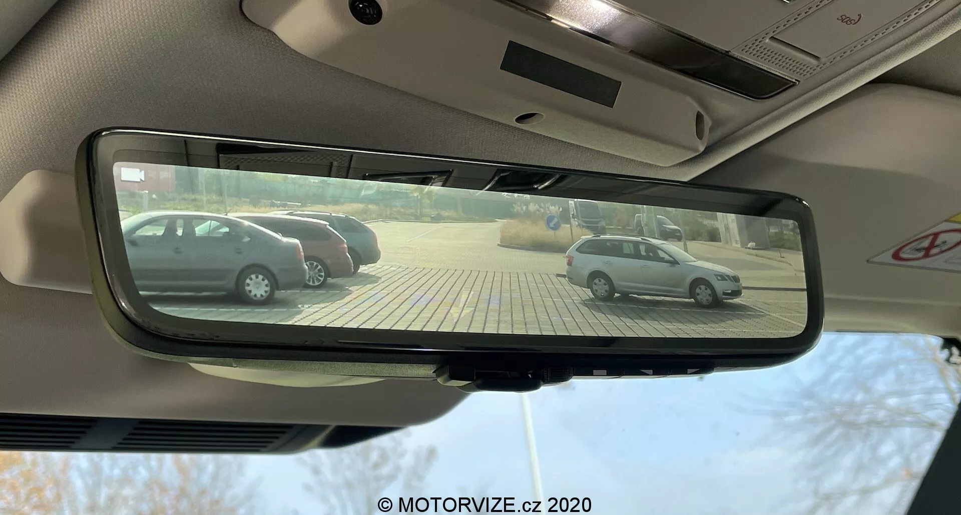 Vista dal sedile del conducente di una Land Rover Defender 2020, con messa a fuoco sullo specchietto retrovisore interno rettangolare auto-oscurante che riflette due hatchback argentate parcheggiate perpendicolarmente in un parcheggio pavimentato con pietre beige in una giornata soleggiata.