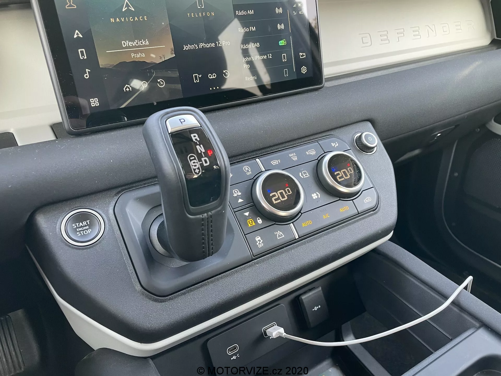 Vista ravvicinata della console centrale della Land Rover Defender 2020 che mette in evidenza la leva del cambio, il sistema di risposta al terreno per varie condizioni di guida, il pulsante di avvio/arresto del motore, i comandi del climatizzatore bi-zona, la porta USB per la connettività del dispositivo, il sistema di infotainment touchscreen parzialmente visibile e i materiali premium utilizzati nel design degli interni, visto dalla prospettiva di un sedile anteriore con un'angolazione di 45 gradi.