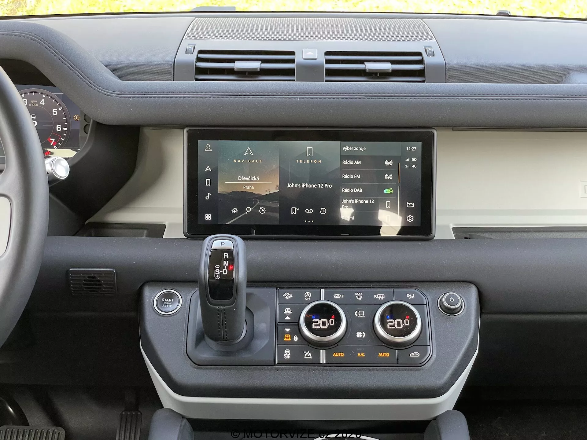 Vista del cruscotto interno del Land Rover Defender 2020 che mostra il sistema di infotainment touchscreen, doppie prese d'aria, selettore del cambio moderno, pannello di controllo del clima a doppia zona, scorcio del volante e una combinazione di plastiche rigide e materiali soft-touch con un contrasto di colore sul cruscotto.