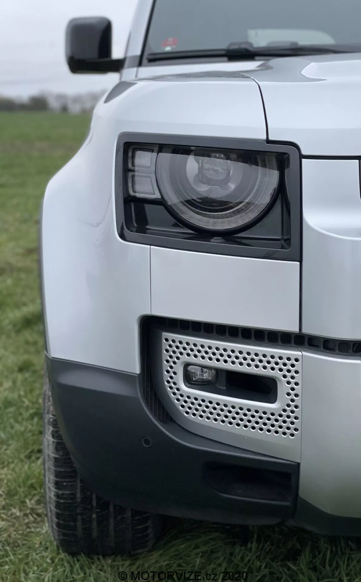 Vista ravvicinata di un Land Rover Defender 2020 con messa a fuoco sul gruppo ottico, griglia e parafango anteriore. L'immagine cattura il veicolo da un'angolazione frontale laterale, evidenziando i moderni fari LED/HID, la tecnologia di illuminazione adattiva, la griglia con possibile posizione dei sensori e la verniciatura bicolore. Il paraurti anteriore mostra una luce antinebbia e un sistema di sensori di parcheggio, enfatizzando il design robusto e contemporaneo del SUV Defender.